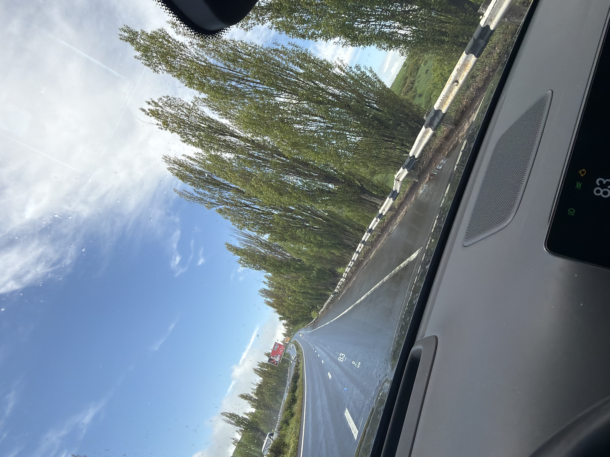 The image shows a view from inside a car driving on a highway. The road is wet, possibly from recent rain, and lined with tall trees. A guardrail runs alongside the road on the right. In the distance, there appears to be a sign or billboard. The sky is blue with some clouds. The car's dashboard is visible in the foreground, with a display showing a temperature of 83 degrees.