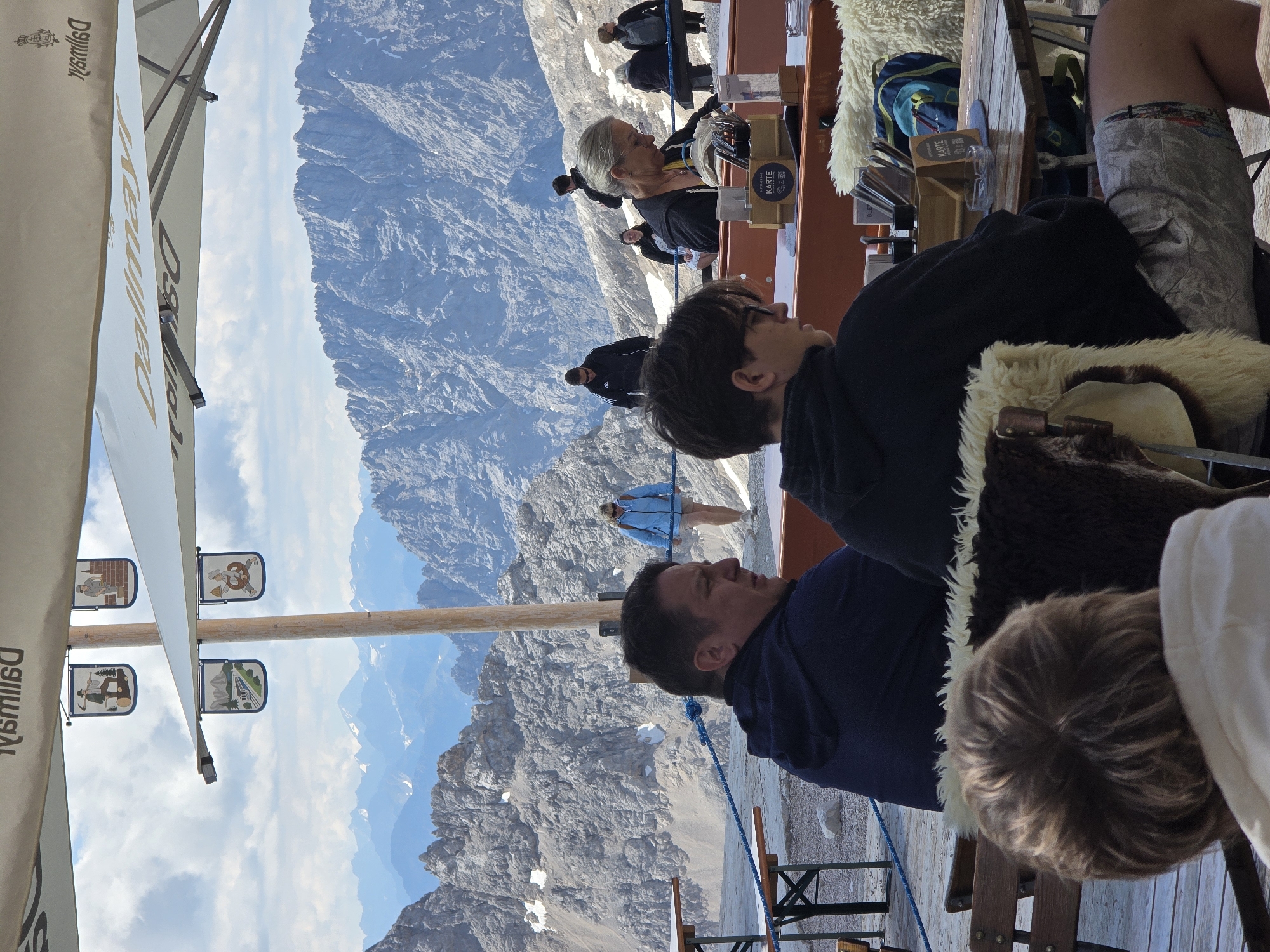 The image captures a scenic mountain restaurant setting, likely on the Zugspitze, Germany's highest peak, near Garmisch-Partenkirchen. The foreground is occupied by people seated at tables, facing away from the camera and towards the stunning mountain panorama. One person in the foreground, with dark, short hair and wearing glasses, is partially visible from the back, as is another person to their left. Further back, a man in a dark blue hoodie is looking to the right, while a woman in a bright blue jacket stands in the middle ground. The background reveals jagged, snow-dusted mountain ranges under a partly cloudy sky. The restaurant area is defined by wooden tables and chairs, some covered with sheepskin throws. Decorative banners depicting traditional scenes hang from a large awning, which is branded with "Dallmayr." A blue rope cordons off the seating area from the rocky terrain beyond. The overall atmosphere suggests a relaxed, outdoor dining experience with breathtaking natural beauty.