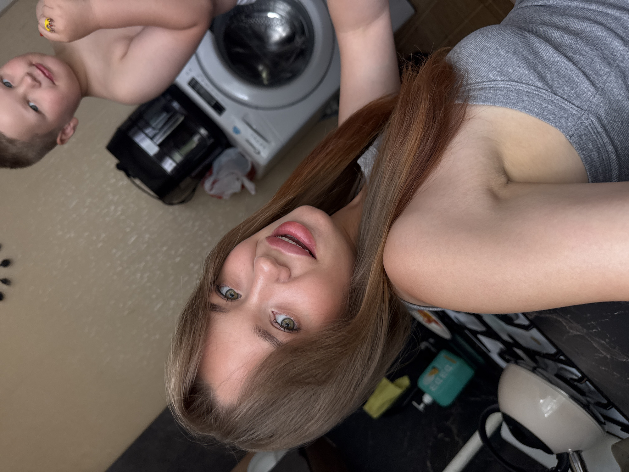 The image shows a young woman with light brown hair and green eyes, smiling at the camera. She is wearing a gray tank top. Behind her, a young boy with short brown hair is visible, looking away from the camera. In the background, there is a washing machine and other kitchen appliances. The scene appears to be in a home kitchen. The city of Chaikovsky, Russia, is not identifiable from the image.