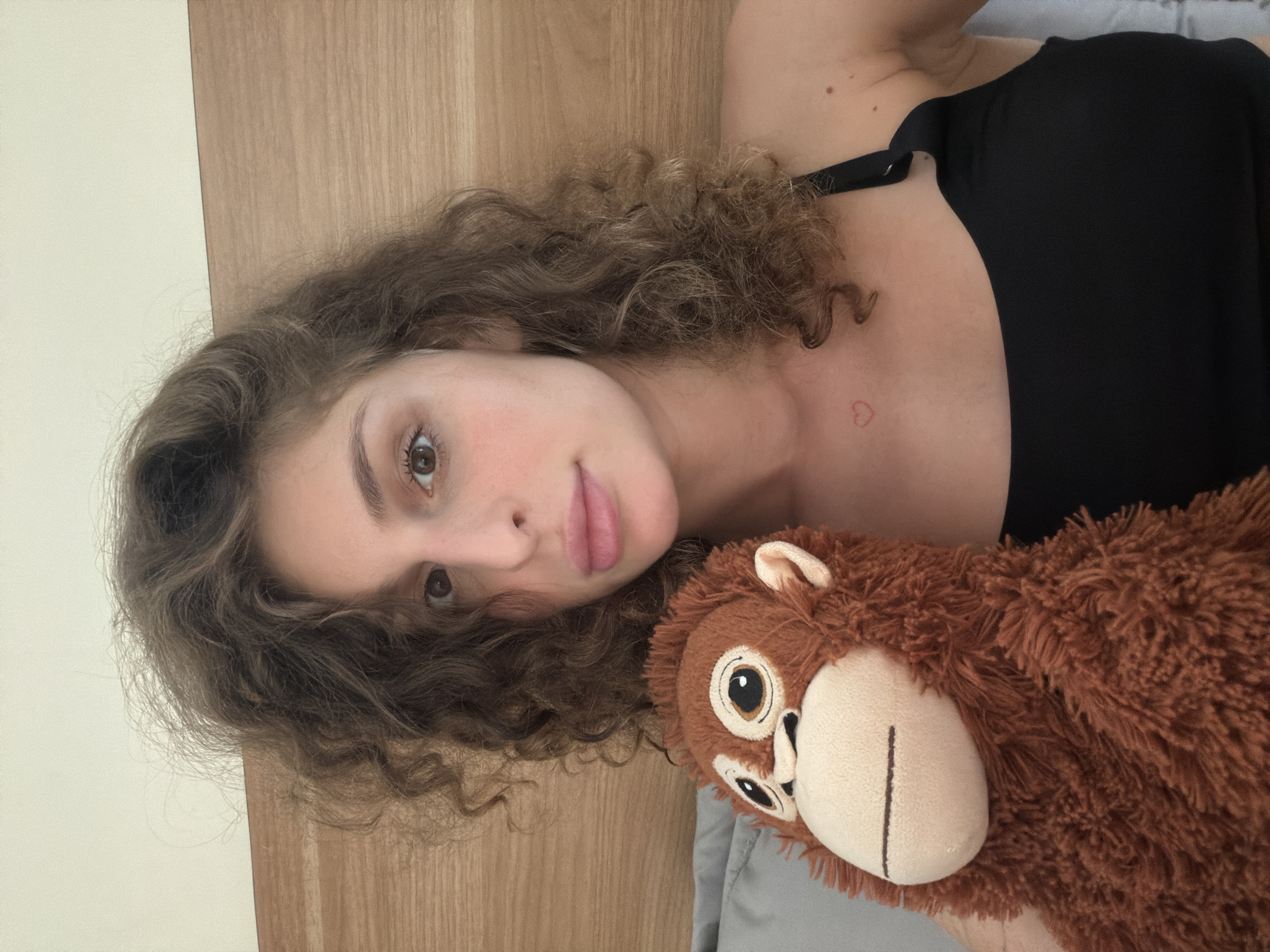 A young woman with curly brown hair and light brown eyes is holding a stuffed monkey. She is wearing a black top. A small red heart tattoo is visible on her upper chest. She is lying in bed, with a wooden headboard behind her. The image appears to have been taken indoors in a bedroom. No specific location in North Kuta, Indonesia can be determined from the image.