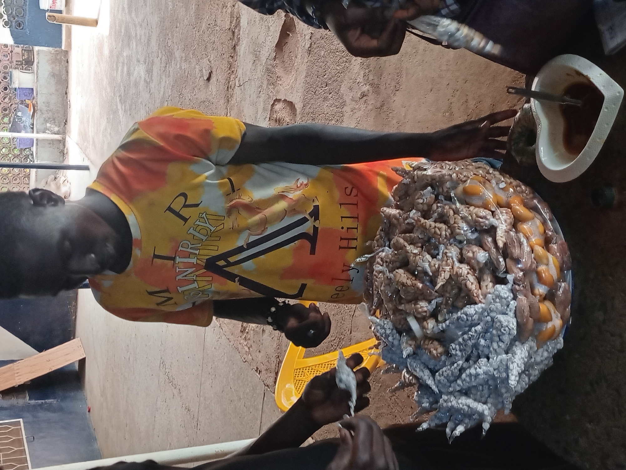The image shows a person in Bukuru, Nigeria, preparing food outdoors. The person is wearing an orange and yellow t-shirt with black text. They are handling what appear to be small octopus, some of which are coated in a white substance. The octopus are piled on a blue plate or platter. Beside the plate is a white, heart-shaped bowl containing a brown sauce, and a metal utensil rests within. The person is using a small, possibly disposable, tool to prepare the food. The background shows an outdoor setting with a mix of rough, unpaved ground and a partially visible building or structure. A yellow plastic basket is also visible nearby.