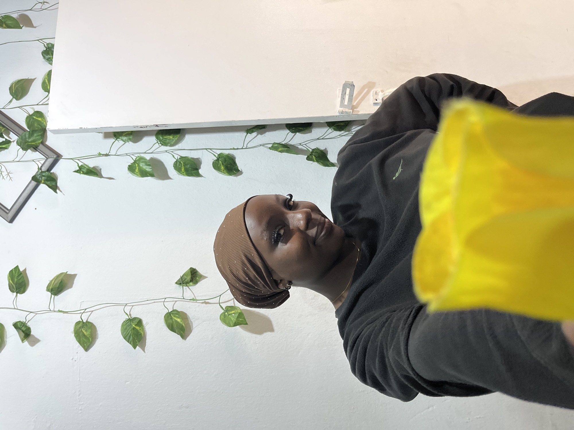 The scene depicts a young woman indoors in Jos, Nigeria, positioned slightly to the left of the frame's center. She has dark skin and is looking directly at the viewer with a subtle smile. She wears a brown, ribbed head covering embellished with small white dots and a dark grey or black long-sleeved fleece sweater featuring a small, light-colored embroidered logo on the left chest. A thin gold-colored necklace is visible around her neck, and she has a small dark earring in her visible left ear. Her left arm extends towards the camera, holding a bright yellow, translucent object, possibly a plastic cup or container, which is significantly out of focus in the foreground.
The background consists of a textured white wall adorned with artificial green leafy vines, some exhibiting yellow variegation, draped vertically. A corner of a grey picture frame is partially visible in the upper left, with vines extending around it. To the right of the woman, a smooth white door or panel is slightly ajar, revealing a metal barrel latch attached to the door frame near its base. Shadows cast by the vines on the wall suggest an interior light source, likely from the right side of the frame.