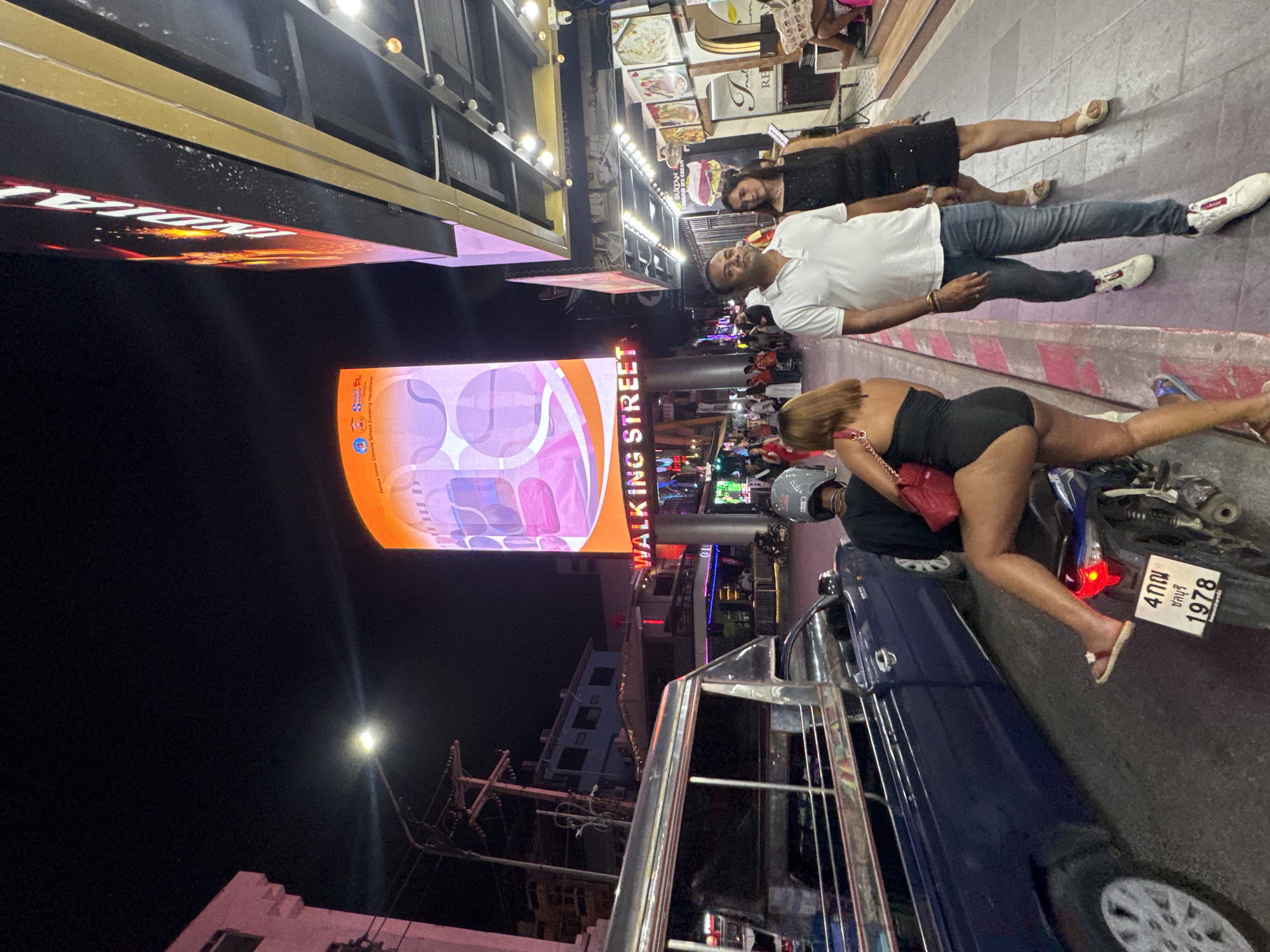 This night scene captures a bustling street in Pattaya City, Thailand, identified by the prominent "WALKING STREET" sign. The time of day is clearly night, with the sky dark and the street illuminated by numerous artificial lights, including neon signs and streetlights, contributing to a vibrant atmosphere.
In the foreground, a woman in a black, form-fitting outfit (possibly a bodysuit or short dress) leans provocatively over a parked motorcycle, holding a red purse. She is positioned next to a dark blue vehicle, possibly a car or a small truck. The motorcycle bears a Thai license plate, reading "4กณ ชลบุรี 1978," confirming the location in Chonburi province. A person wearing a helmet is visible on the motorcycle.
Further back, a man in a white polo shirt and jeans stands alongside a woman in a black dress; both are looking towards the viewer, seemingly observing the scene or posing for a photo. The background reveals a lively street filled with more people, suggesting a crowded pedestrian area, flanked by buildings with illuminated storefronts. Electrical poles and overhead wires are also visible against the dark sky.
Visible text includes the large "WALKING STREET" sign, which also displays "smart Percaya Senior Citizens School Creating Happiness" and "SULTAN TURKISH ICE CREAM" on a smaller sign. The overall scene depicts a typical busy night in a well-known entertainment district.