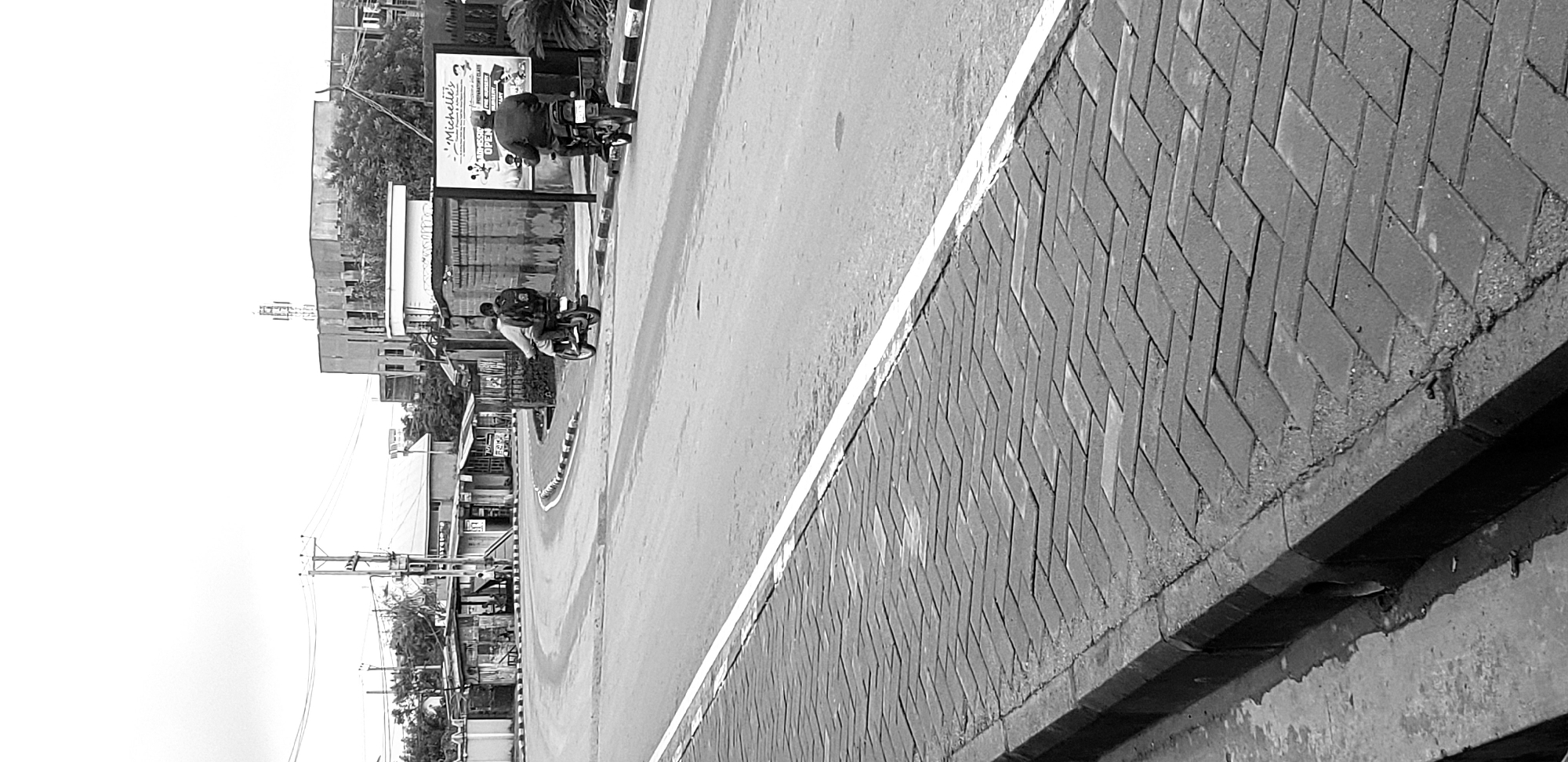 A black and white, eye-level, medium shot captures a street scene in Narayi, Nigeria. The foreground is dominated by a herringbone pattern of grey brick paving stones that form a sidewalk, bordered by a concrete curb. A white line divides this sidewalk from the asphalt road, which curves to the left and recedes into the background. Two individuals on motorcycles are the primary subjects. The motorcycle on the left carries a person with a backpack, both facing away from the camera and traveling in the direction of the road's curve. The motorcycle on the right has a rider also facing away from the camera, moving in the same direction. To the right of the road, a billboard with text and images, including the word "Michelle's," stands next to a brick wall. In the background, a series of buildings, some appearing commercial with awnings, are visible, along with utility poles and trees. The sky is overcast, suggesting a cloudy day.