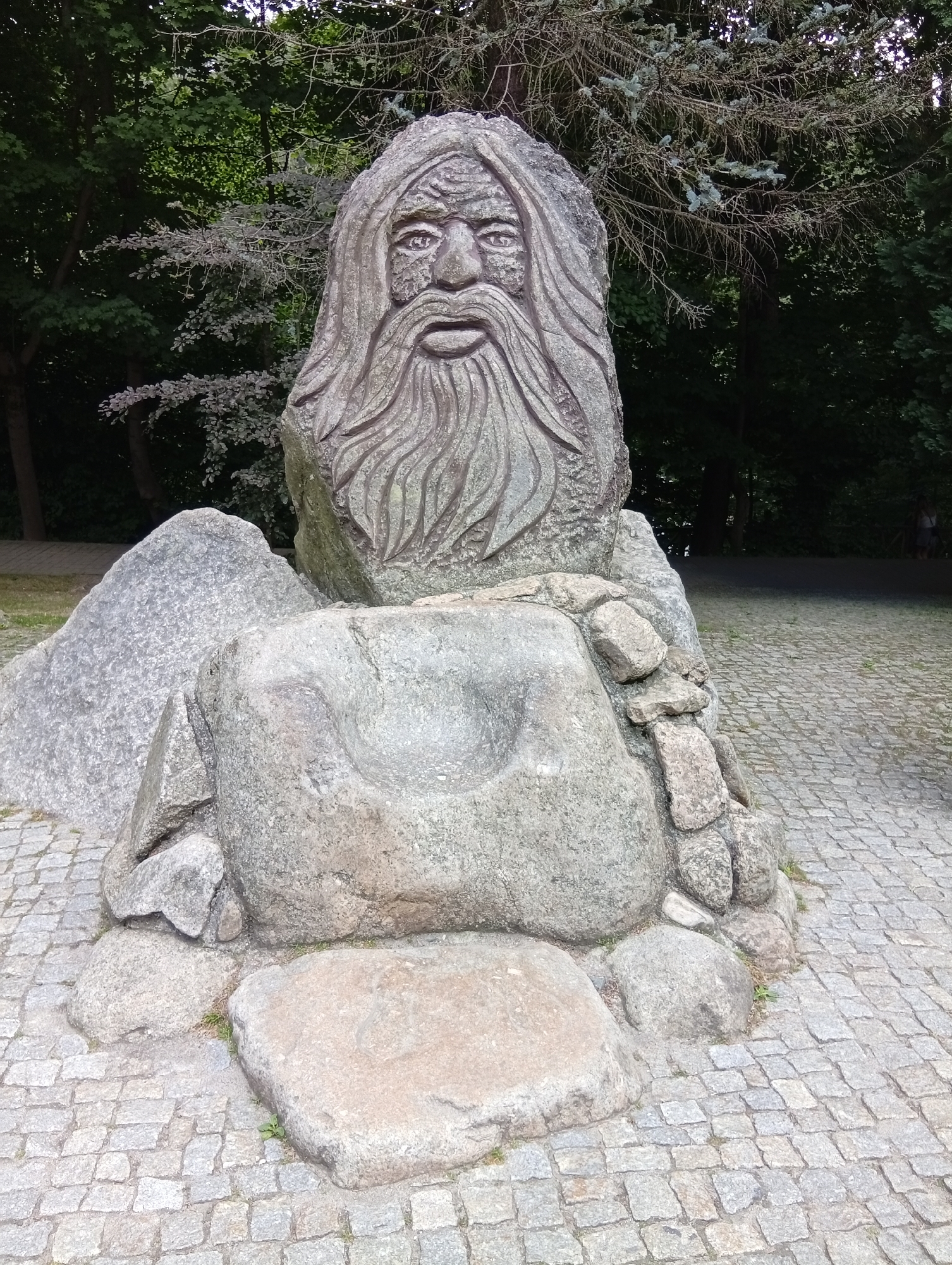 This is a full-shot, eye-level, outdoor photograph of a large, carved stone sculpture of a bearded man's face. The sculpture is situated in a park-like setting with cobblestone paving in the foreground and trees in the background. The sculpture is composed of several large, rough stones, with the carved face being the prominent feature. The face is depicted with deep-set eyes, a wide mouth, and flowing hair and beard. The carving is detailed, showing textures that mimic skin and hair. In front of the main carved stone, there is a large, roughly carved stone chair or seat. The surrounding stones appear natural and are of various sizes and shapes. The cobblestone pavement is laid in a uniform pattern. The background consists of dense green trees, suggesting a wooded area or park. Sunlight filters through the trees, creating some dappled light and shadow. There is no discernible human activity or interaction in the immediate vicinity of the sculpture, though a faint figure of a person can be seen in the far right background. This sculpture is known as the "Stone Giant" and is a landmark located in Karpacz, Poland.
