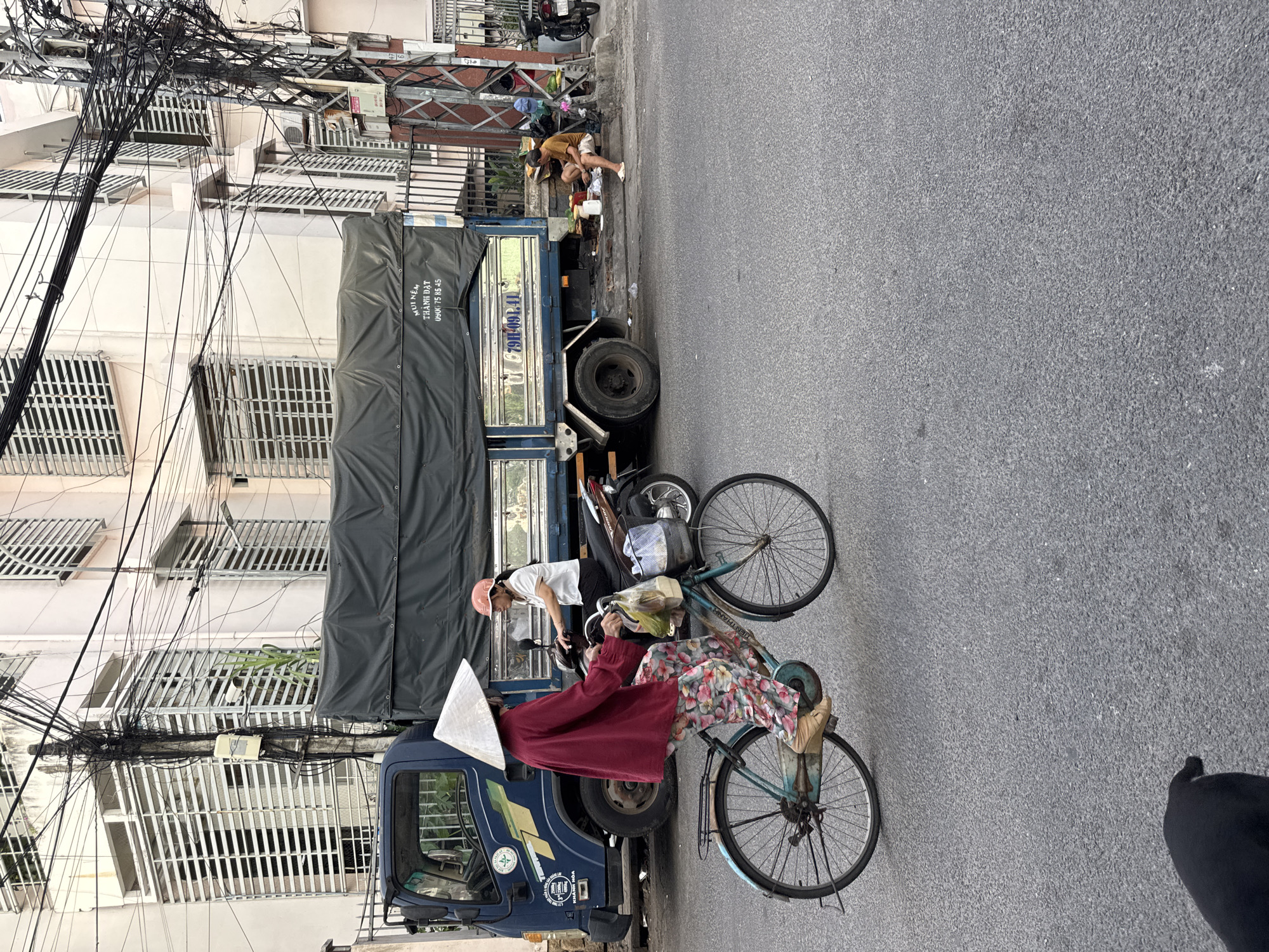 In Nha Trang, Vietnam, on a paved street, a woman wearing a traditional conical hat and a vibrant floral patterned outfit is riding a bicycle. She is dressed in a loose red long-sleeved shirt and light-colored trousers adorned with colorful flowers and leaves. Her feet are clad in beige wedge shoes. The bicycle is a light blue color with a basket in the front, carrying plastic bags containing groceries. Behind her, a person on a scooter, wearing a white shirt and a pink helmet, is also on the street.
A large blue truck with a dark tarp covering its cargo bed is parked on the right side of the road. The truck has some text and a license plate visible. Near the truck, a person in a brown shirt and white shorts is sitting on the curb, engaged in some activity with items laid out in front of them, suggesting a small street-side setup, possibly for selling goods or preparing food.
The environment includes a building with white horizontal blinds on its windows in the background, and a complex network of electrical wires suspended above the street, indicative of urban infrastructure. The street is asphalt, and the overall scene depicts a typical day in a Vietnamese city with its characteristic blend of vehicles, people, and bustling street life.