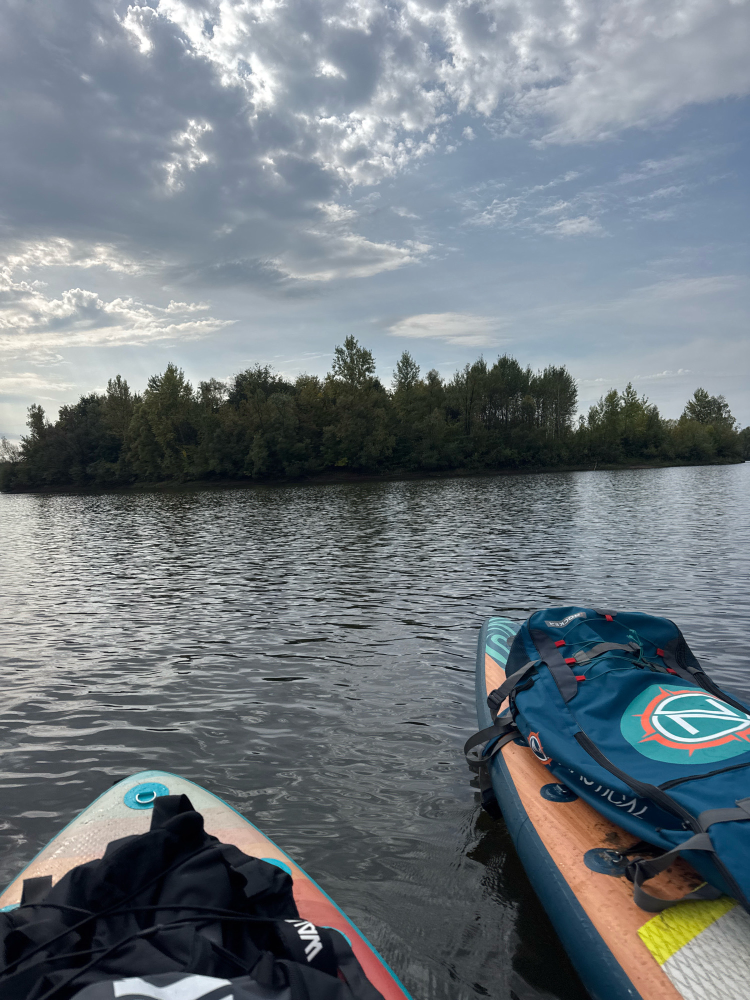 The image depicts a serene view from a stand-up paddleboard on a body of water. In the foreground, two paddleboards are visible, each with gear strapped to it. One paddleboard has a dark bag and a black jacket, while the other has a teal duffel bag with a circular logo. The water is calm with ripples reflecting the cloudy sky. In the background, a dense line of trees forms a treeline along the distant shore, with some taller trees interspersed. The sky is overcast with dramatic clouds, suggesting it may be early morning or late afternoon. The location is identified as the Belaya River in Ufa, Russia, a common spot for recreational water activities.