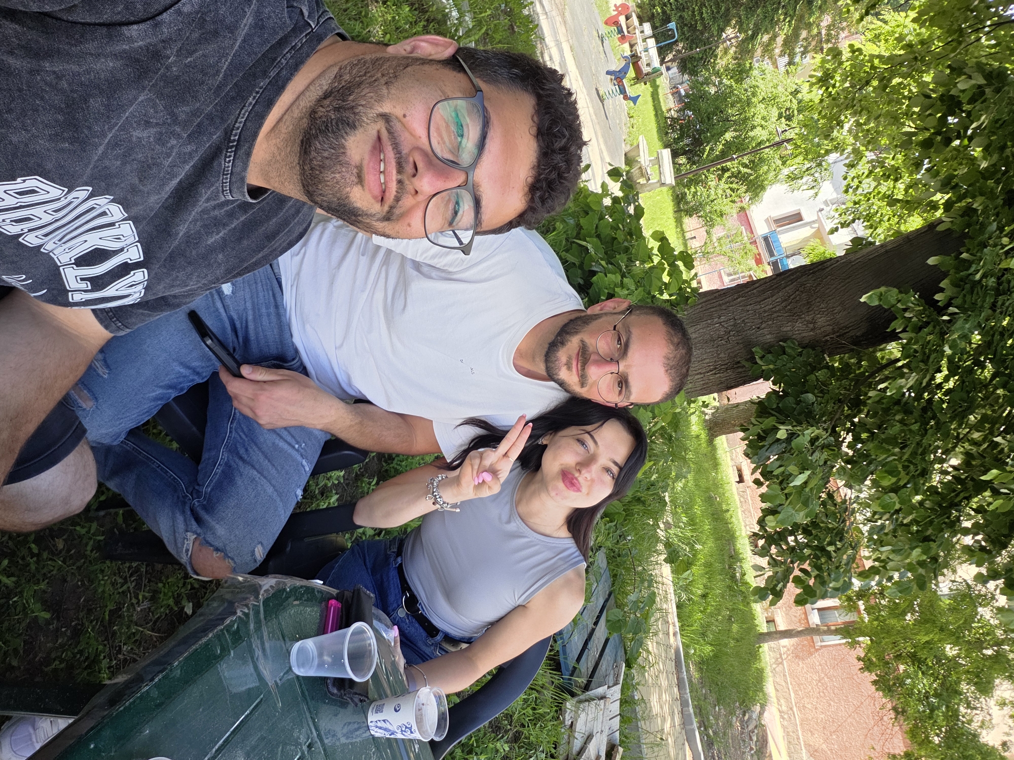 Three individuals are posing for a selfie outdoors. One individual is wearing glasses and a dark t-shirt, and another is wearing a white t-shirt and glasses. A woman is sitting at a table and is making a peace sign with her fingers. The table has multiple plastic cups on it. The setting appears to be a park or garden in the city of Rudozem, Bulgaria, with trees and grass visible in the background. A building and playground are also in the background.