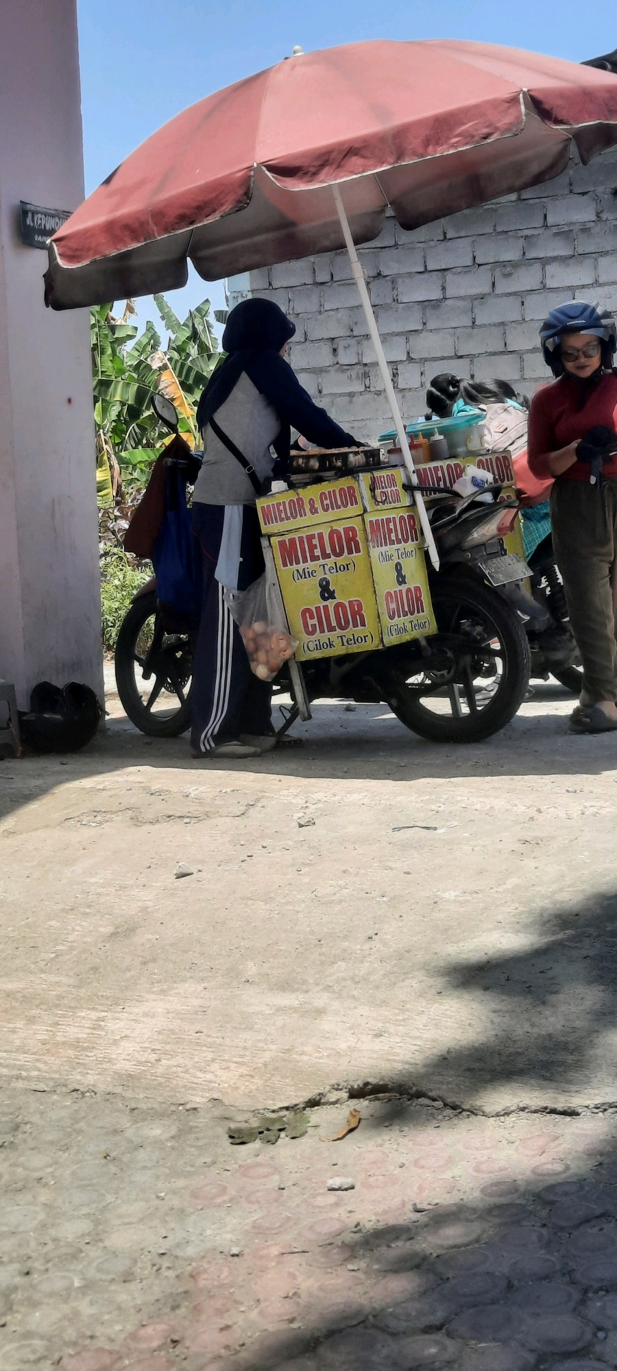 In Tangkil, Indonesia, a person wearing a dark blue hoodie and gray shirt, along with white and black striped pants and sandals, stands by a motorcycle-mounted food cart. The cart, with yellow signage that reads "MIELOR & CILOR (Mie Telor) & CILOR (Cilok Telor)," displays several packages of eggs in a clear plastic bag. Another person, wearing a red shirt, brown pants, and a blue helmet, stands to the right, holding a black object. A large red umbrella shades the food cart. The scene unfolds outdoors on a paved surface, with a brick wall and lush green foliage in the background. A street sign reading "JL. KEPUNDUNG" is visible on the left. The lighting suggests it is daytime.