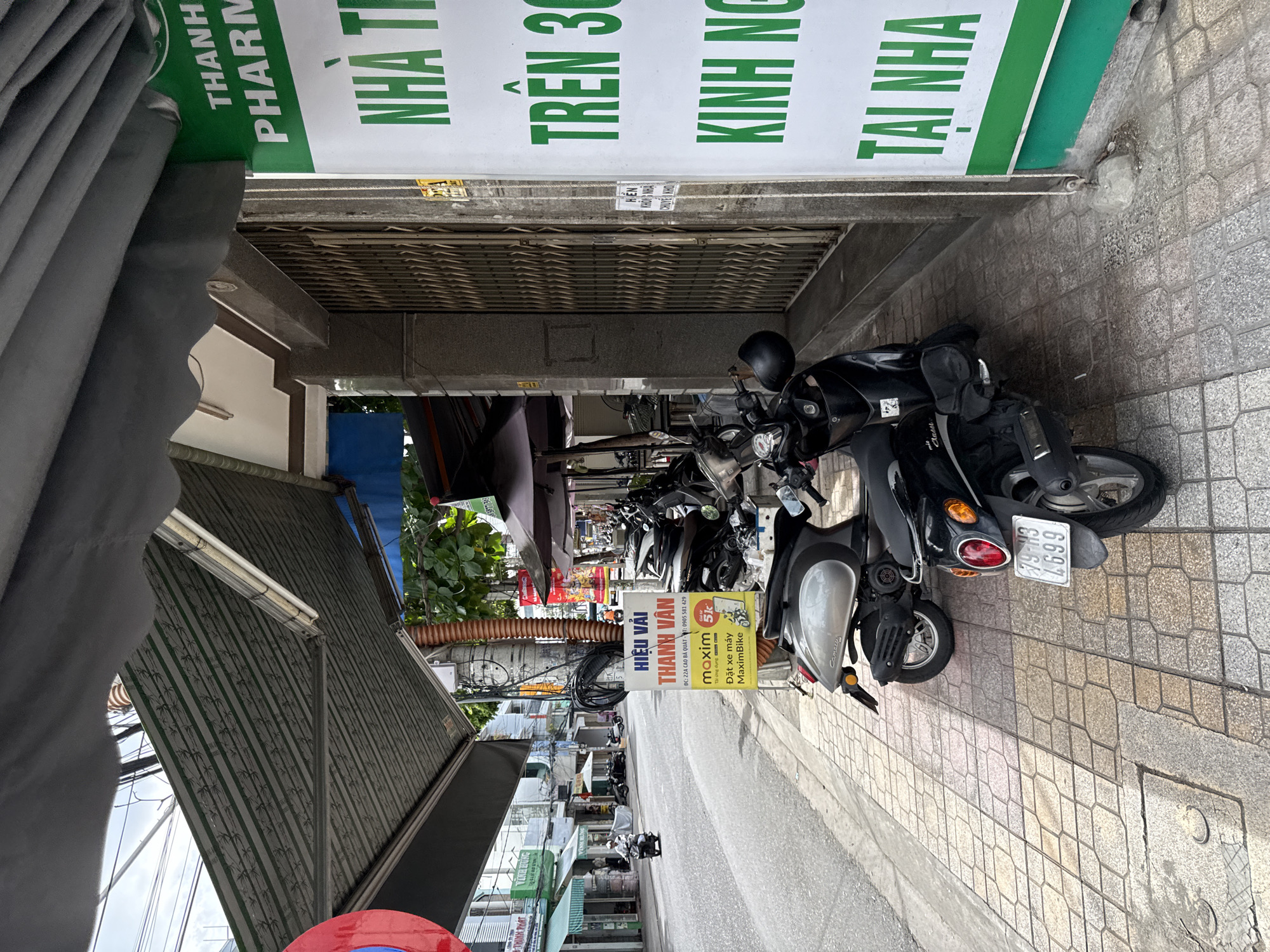 The image depicts a street scene in Nha Trang, Vietnam. In the foreground, several parked motorbikes are visible on a paved sidewalk. A light-colored scooter is on the left, and a black scooter is on the right with its license plate reading "79-H3 4699". A sign with green text on a white background is attached to a pole, displaying Vietnamese writing. Behind the motorbikes, the street continues with more parked vehicles and buildings lining the side of the road. An awning extends over the sidewalk from one of the buildings. The sky is overcast.
