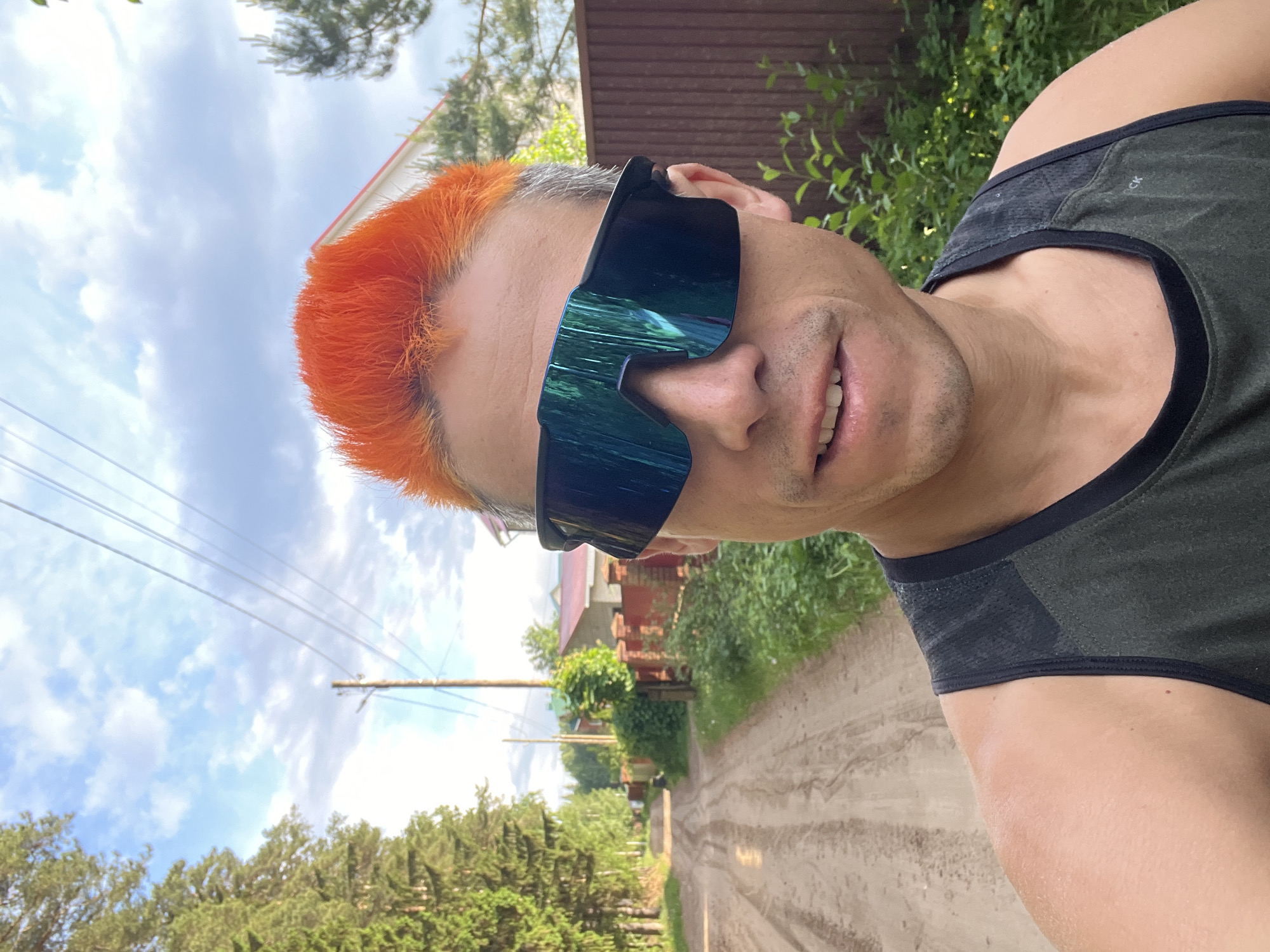 A selfie was taken in Chaikovsky, Russia. The subject, a man with short orange hair and wearing dark, reflective sunglasses, is partially framed in the upper right. He also wears a dark tank top. The background shows a dirt road lined with green foliage and trees. Overhead are utility poles and power lines. In the distance are houses with red roofs. The sky is visible through the trees and is partly cloudy.