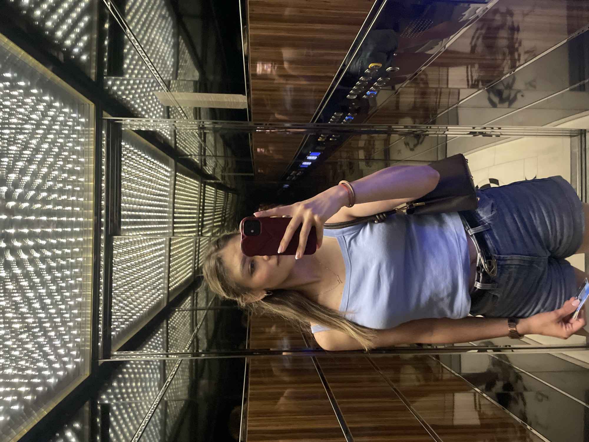 A young woman with blonde hair tied in a ponytail takes a mirror selfie in an elevator. She is wearing a light blue tank top, denim shorts, and a black belt with a silver buckle. She has a dark handbag slung over her left shoulder and a smartwatch on her left wrist. In her right hand, she holds a smartphone with a burgundy case, using it to take the photo. The elevator is lined with reflective surfaces and features a ceiling with a repetitive pattern of lights. The walls are paneled with wood-grain laminate. The floor is tiled in a checkered pattern. The overall impression is one of a modern, well-lit interior space.