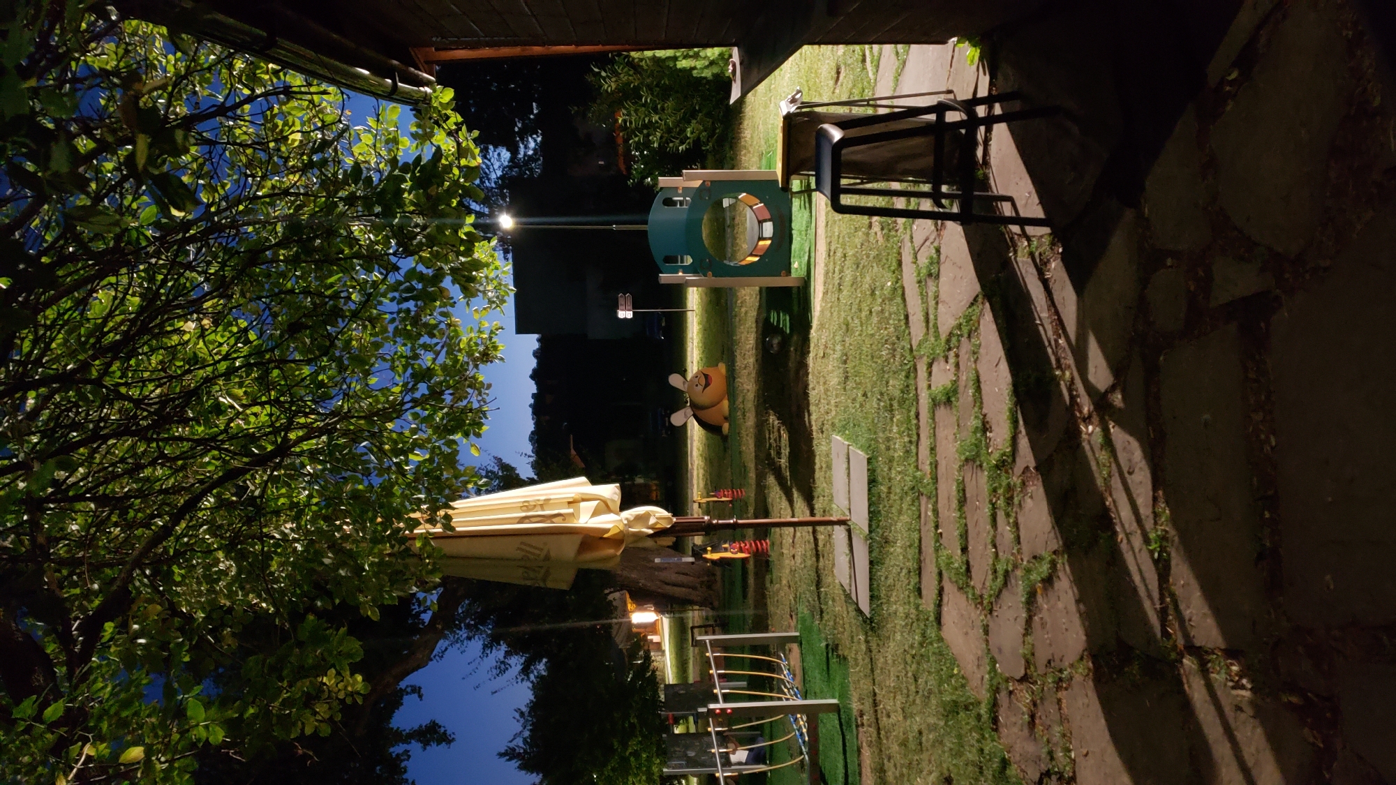 The image depicts an outdoor children's playground at dusk or night, illuminated by artificial lighting. The playground is situated in a grassy area with stone paving in the foreground. Key features include a large, partially closed yellow umbrella, a green and teal climbing structure with a circular opening, and a bumblebee-shaped spring rider. A bar stool is visible in the foreground, casting a long shadow. In the background, trees line the perimeter of the playground, and a building with a dark facade is partially visible on the right. The sky is a deep blue, indicating twilight or a clear night. The location is identified as Prešov, Slovakia. There are no people visible in the image.