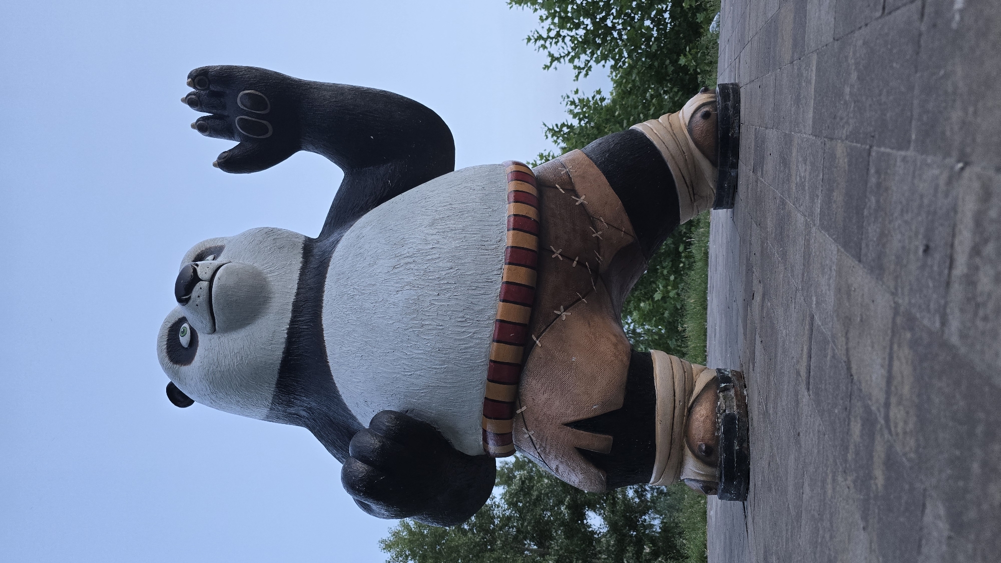 A low-angle, full-body outdoor photograph depicts a large, full-color statue of the animated character Po from Kung Fu Panda, located in the city of Kyiv, Ukraine. The statue features a panda character with black and white coloration, green eyes, a black nose, and an upward-curved mouth. Its left arm is bent at the elbow with the hand clenched into a fist, while its right arm is raised with an open palm, fingers extended. The character wears reddish-tan colored shorts with visible lighter-colored cross-stitching, a prominent red and yellow horizontally striped belt or sash around its waist, and light-colored bandages wrapped around its lower legs and ankles, ending in dark, block-like footwear bases. The statue stands on a ground surface paved with gray, rectangular interlocking pavers or tiles, which are visible from the bottom foreground up to the statue's feet. The background consists of a clear, light blue sky in the upper portion of the frame, with green deciduous trees and foliage visible behind the statue on both the left and right sides. The lighting is diffuse and even, indicating an outdoor setting, possibly during an overcast day or twilight hours.