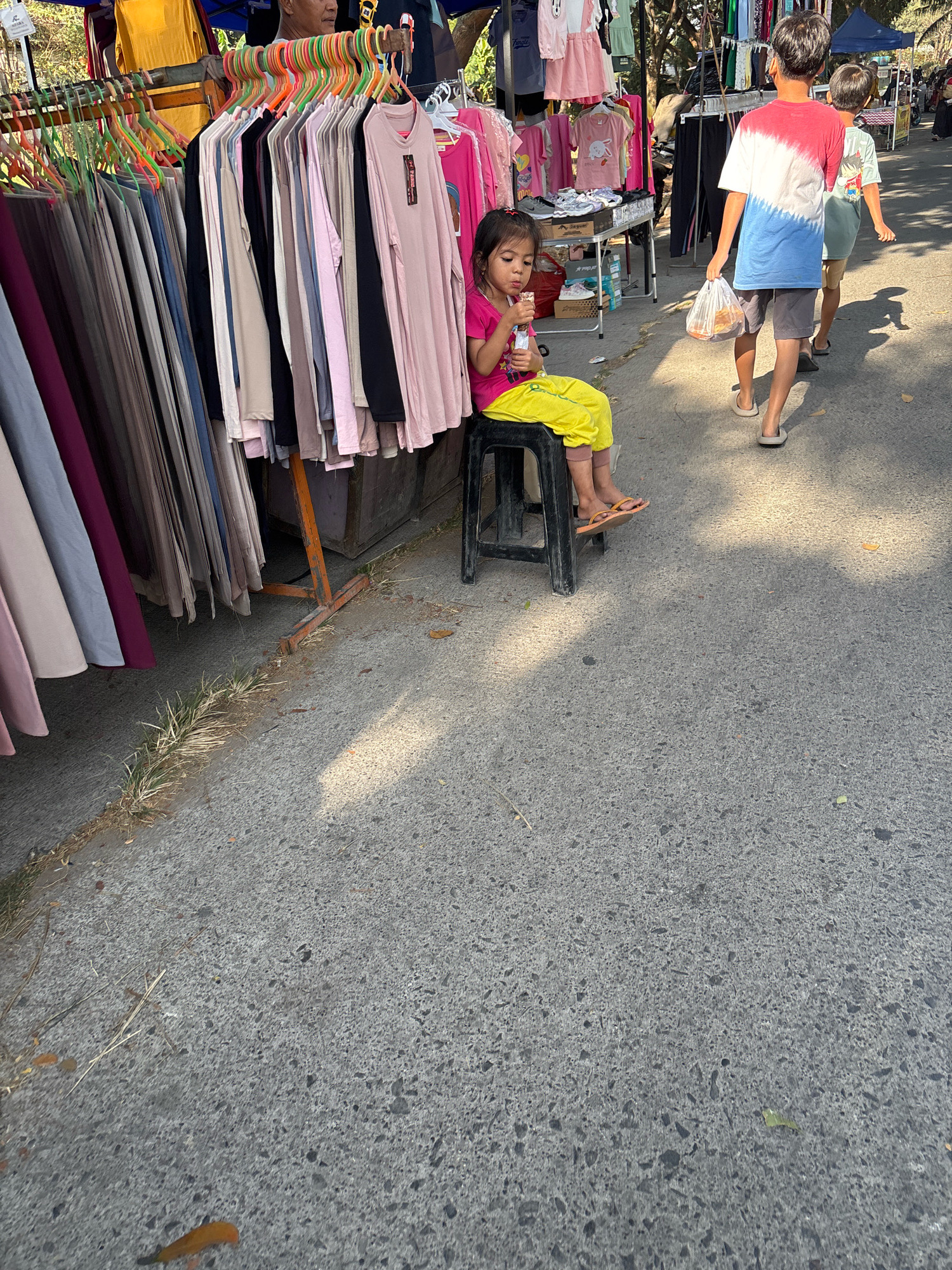 A young girl wearing a pink shirt and yellow pants sits on a black plastic stool at a street market in Telagamurni, Indonesia. She is eating from a package with a utensil. In the background, two young boys walk away from the camera, the one in front carrying a plastic bag. To the left, racks of colorful clothing are displayed for sale. The market is set up along a paved street, with trees and other stalls visible in the distance. The lighting suggests it is a sunny day, with shadows cast across the ground.