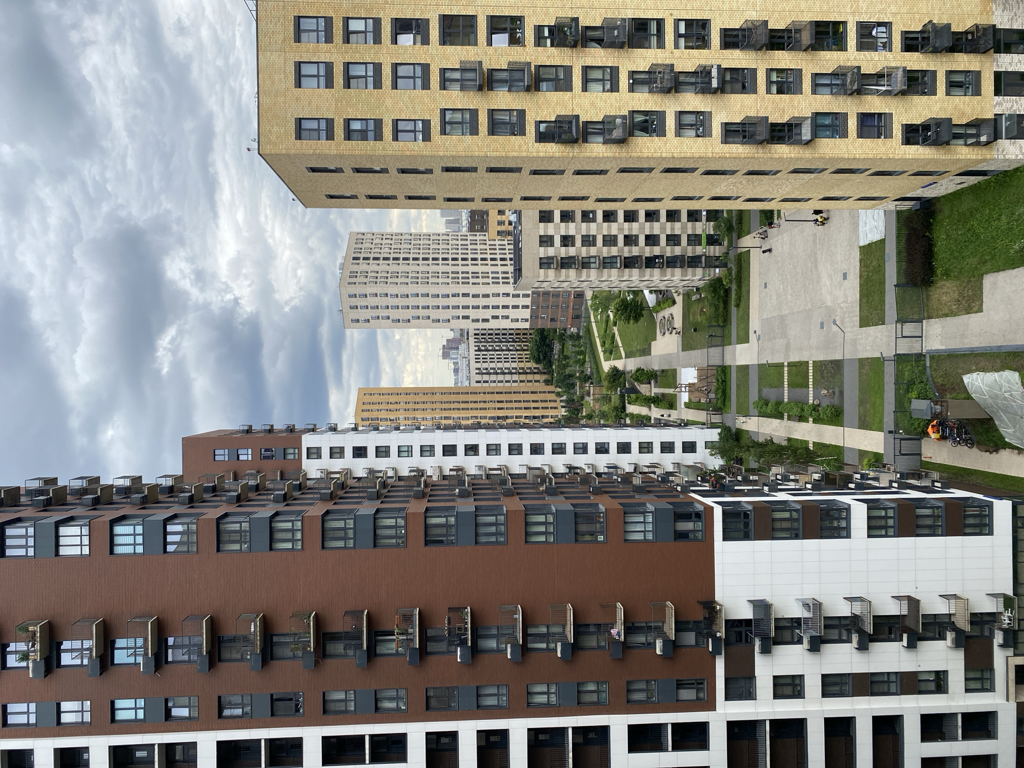 An elevated, direct, eye-level perspective shot captures a view of a modern residential complex in Moscow, Russia. The sky above is overcast, filled with dramatic, heavy grey clouds. Dominating the foreground are several tall apartment buildings, characterized by their rectangular structures, varied facade colors, and numerous balconies.
On the left, a predominantly white building stands tall, its numerous windows and balconies creating a repeating pattern. Adjacent to it, a building with a rich, reddish-brown brick facade rises, also featuring rows of windows and dark-railed balconies. Further to the right, a tall, light yellow building with a similar pattern of windows and balconies is prominent. In the middle ground, other apartment buildings of varying heights and colors are visible, including a tall, light-colored building with a distinctive grid-like pattern of windows.
Below the buildings, a courtyard area is laid out with paved pathways, grassy lawns, and some playground equipment, including swings and slides. A few people are scattered throughout the courtyard, with some appearing to be walking or gathered in small groups. On one of the paved areas, two individuals are visible near some parked bicycles and an orange tarp. The overall scene suggests a contemporary urban living environment with extensive green spaces incorporated into the design.