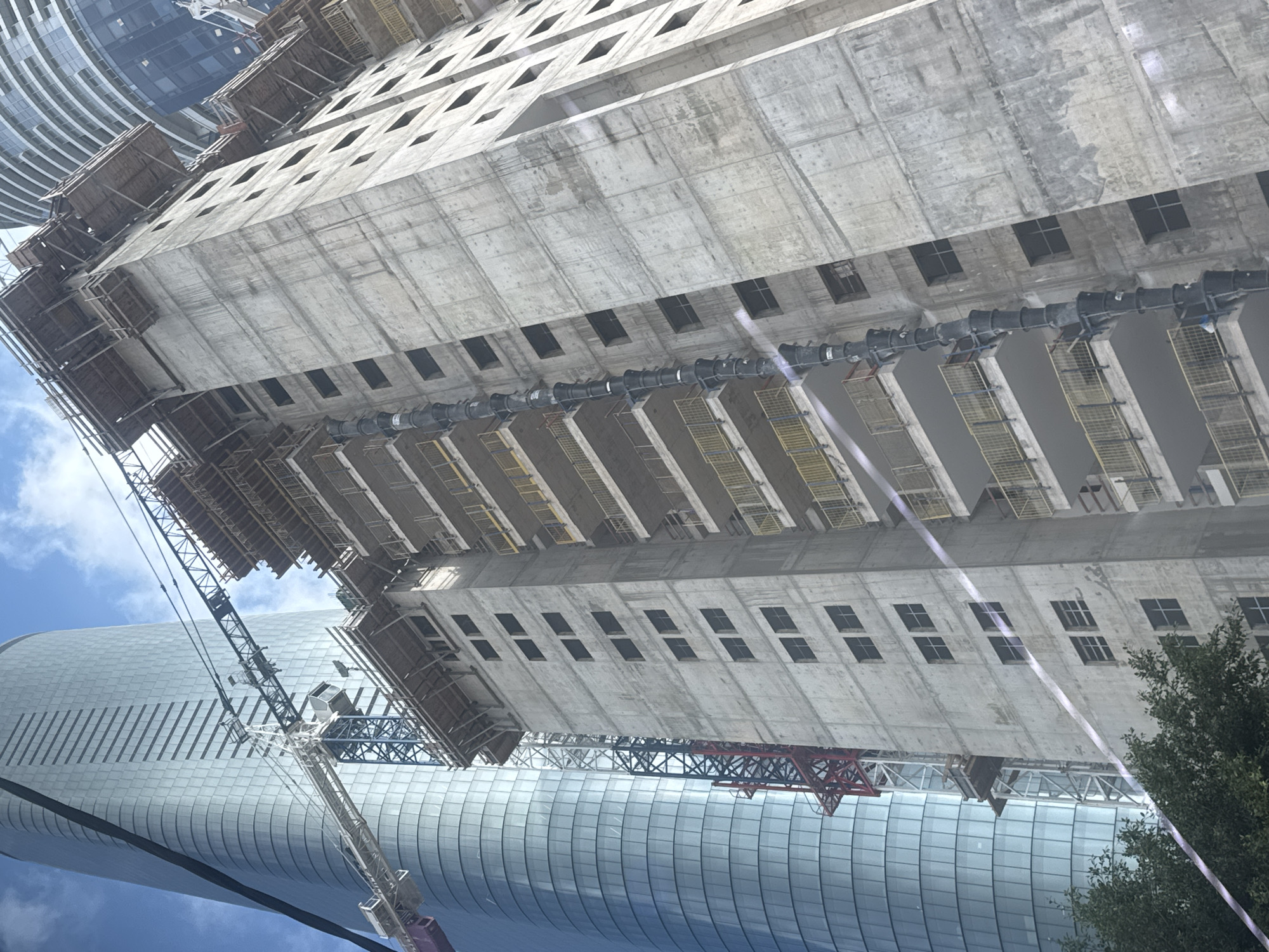 The image shows a construction site in Givatayim, Israel. A tall, rectangular concrete building under construction dominates the view. Its facade has rows of square window openings. Construction scaffolding and a crane are visible. A modern, curved skyscraper with a blue-tinted glass facade is in the background. Part of a tree is on the right. The sky is blue with scattered clouds. A large black corrugated air-conditioning duct is strung on the side of the construction project.