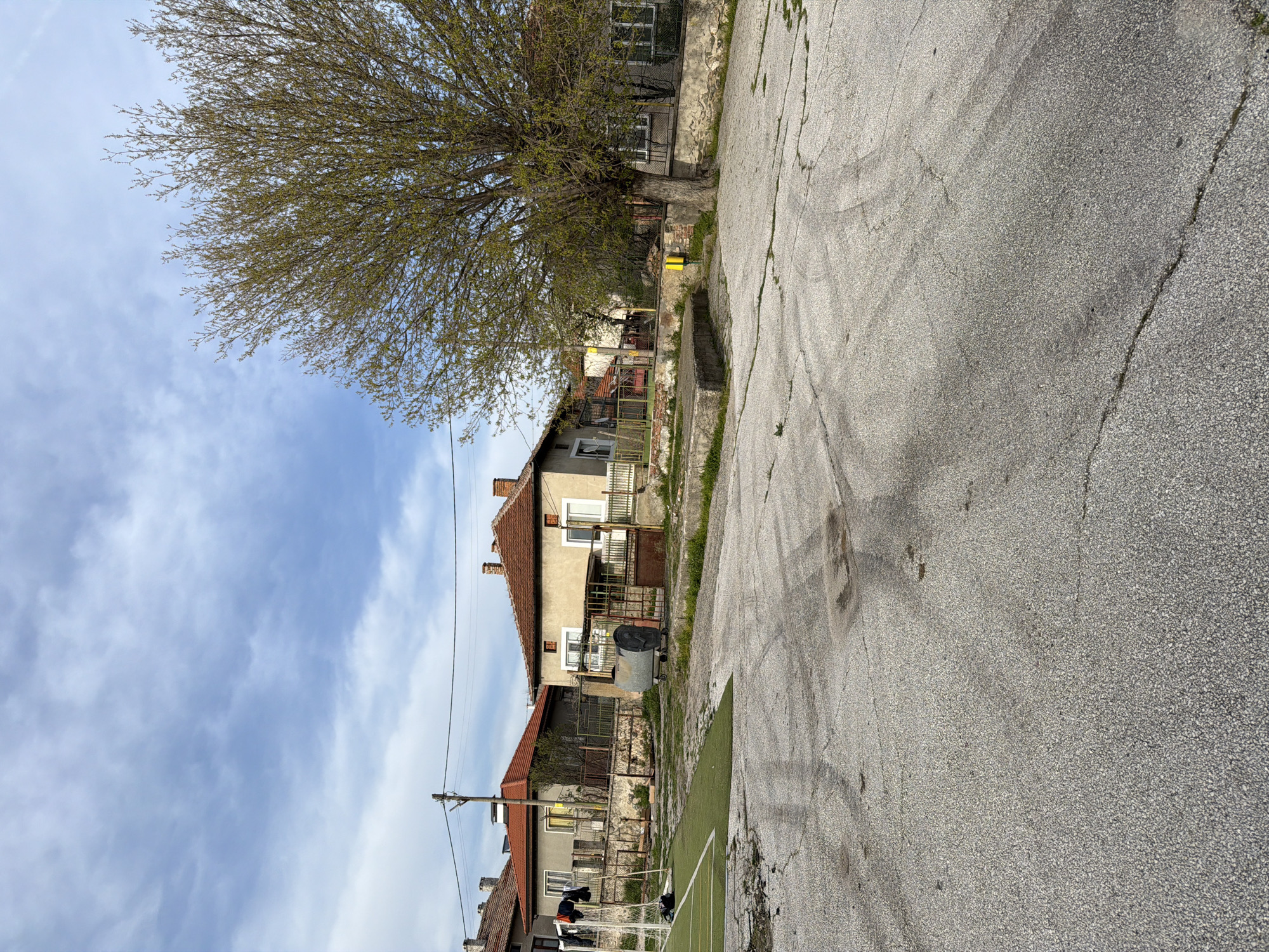 The image shows a street scene in Popintsi, Bulgaria. The street is paved with cracked asphalt. A large tree stands on the left, partially obscuring a building with a red-tiled roof and a balcony. Another building is visible behind it. Power lines run along the side of the street. The sky is blue with scattered clouds. A soccer field with white lines is visible near the bottom of the image. A person is walking near a soccer net.