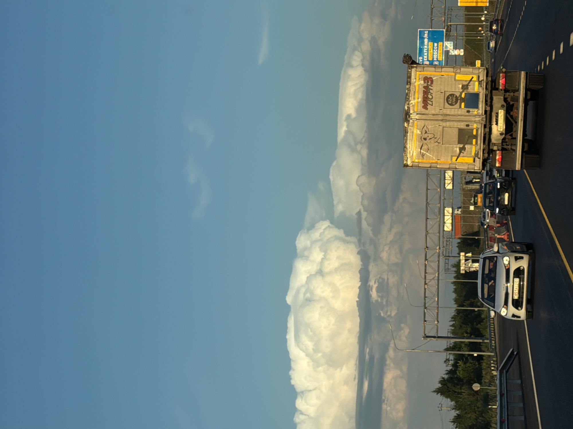 The image captures a sunny day in Egor'evo, Russia, with a clear blue sky dominating the upper portion and dramatic, large cumulonimbus clouds visible on the horizon. The scene is set on a multi-lane highway, with several vehicles in motion. In the foreground, a white sedan is traveling in the left lane, and a large white and yellow refrigerated truck is in the right lane. The truck has "MEGA LIGHT 3" written on its side and a drawing of a bull. In the background, other cars and trucks are visible, along with highway infrastructure including overhead signs and light poles. Trees line the sides of the highway. One blue highway sign in the distance indicates directions to Yekaterinburg, Moscow, and Tolyatti. The overall impression is one of a typical highway scene under a dynamic sky.