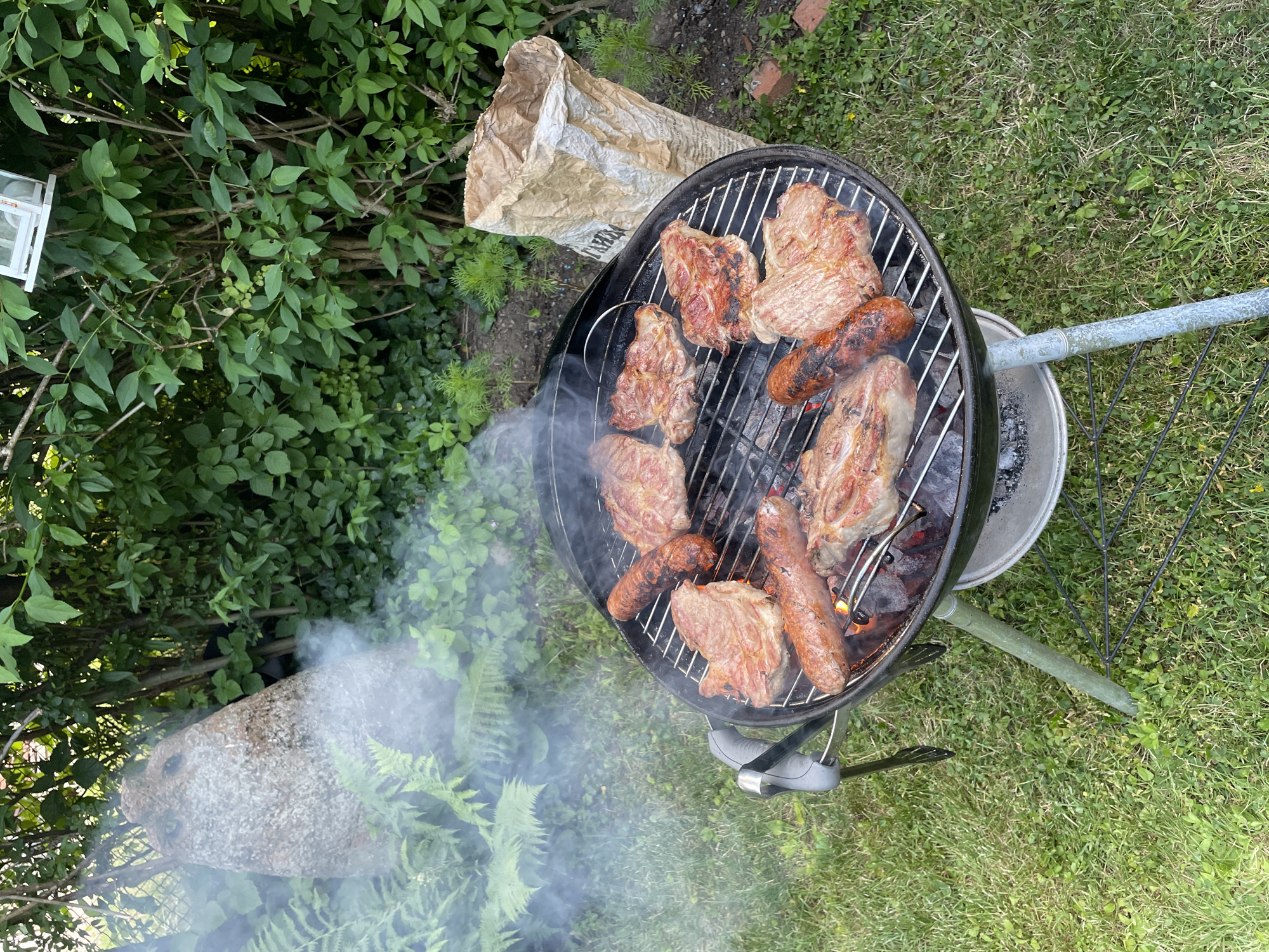 A round charcoal grill is set up on a grassy lawn, with meat and sausages cooking on its grate. Smoke rises from the grill, obscuring some of the background. In the background, there is lush green foliage, a large stone with a hole in it, and a white paper bag that appears to contain charcoal. The scene suggests an outdoor barbecue in a garden setting. The location is not specified as Sofia, Bulgaria.