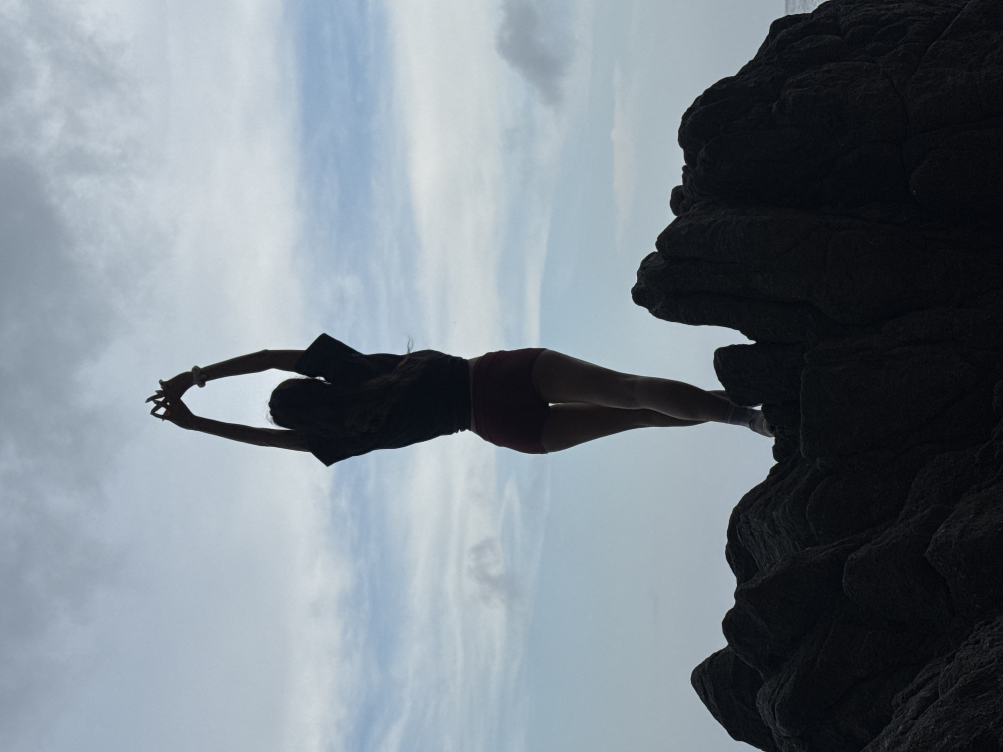 A silhouette of a woman stretching against a cloudy sky background, standing on a rock formation that seems to be a part of the cliffs at the Son Tra Peninsula in Đà Nẵng, Vietnam. The woman is wearing what looks to be a dark-colored t-shirt and red shorts and is reaching upwards with her arms outstretched.