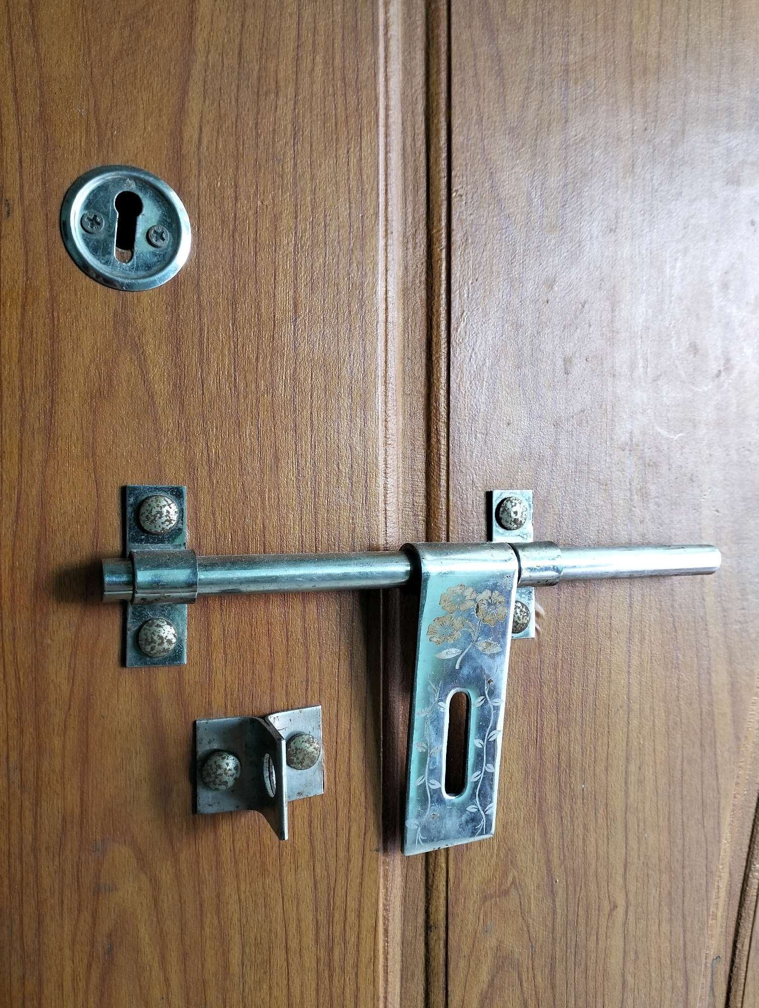 The image displays a close-up view of a wooden door with a metal latch and a keyhole. The door has a visible wood grain texture. Mounted on the door is a silver-toned metal sliding bolt latch, attached with screws to two metal brackets. One bracket is on the main part of the door, and the other is on the edge of the door, designed to receive the bolt. Above the latch mechanism, there is a circular metal keyhole plate with a keyhole opening. The latch plate, which the bolt slides into, has an etched floral design. Another smaller metal bracket, also with two screws, is mounted below the main latch bracket. The scene is set in Sriperumbudur, India, and the overall impression is of a functional, albeit somewhat worn, door security system.