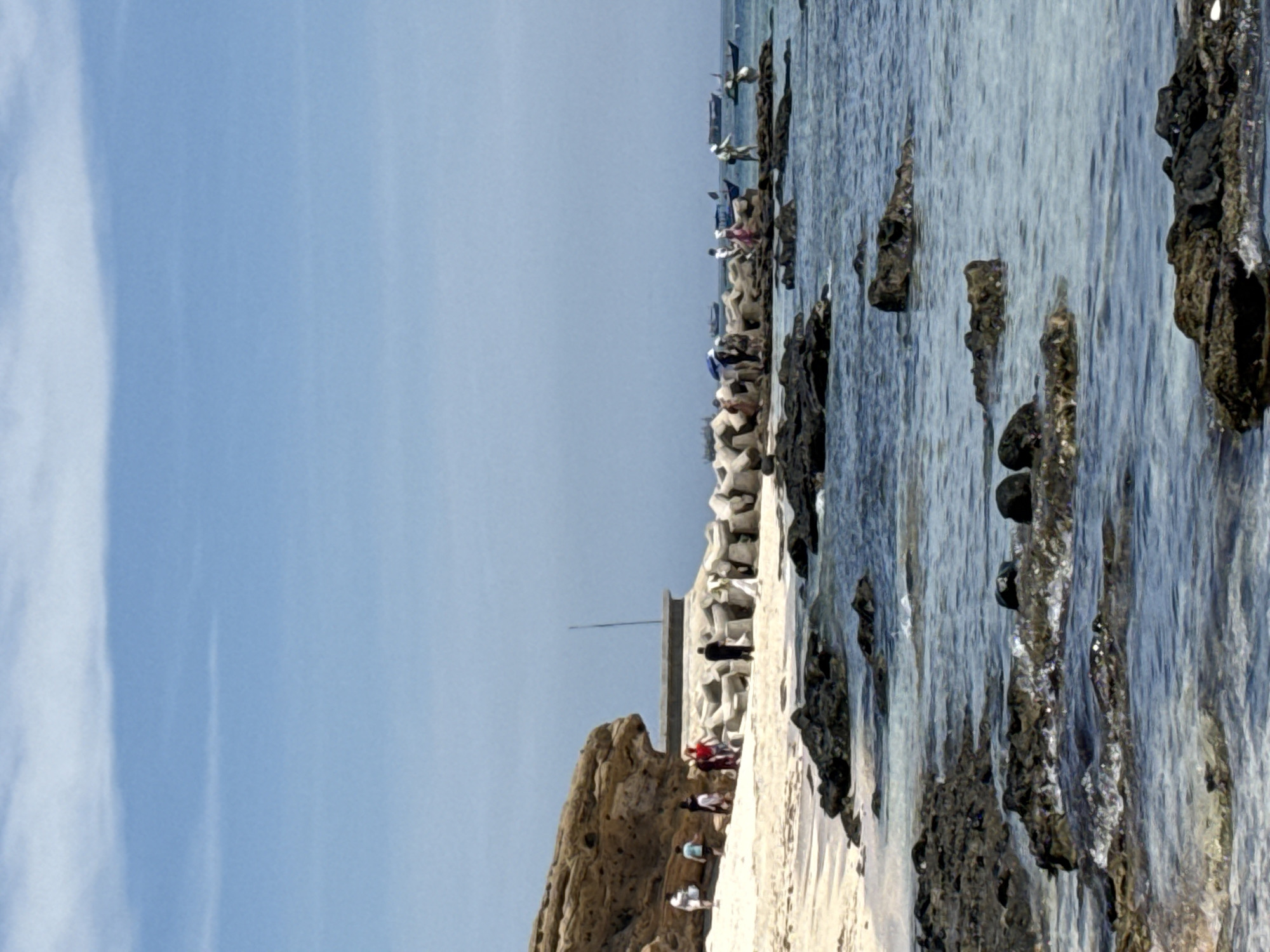 The image depicts a coastal scene in Lý Sơn Special Administrative Region, Vietnam, featuring a rocky shoreline with the sea on the right and a sandy beach on the left. Several people are visible in the mid-ground, walking along the beach and near a breakwater constructed from concrete tetrapods. Some individuals are gathered around a small building. In the background, several boats are anchored in the water. The water is clear and calm, with small waves lapping against the rocks. The sky is a clear blue with a few wispy clouds. The overall impression is one of a tourist destination, possibly a fishing village, on a sunny day.