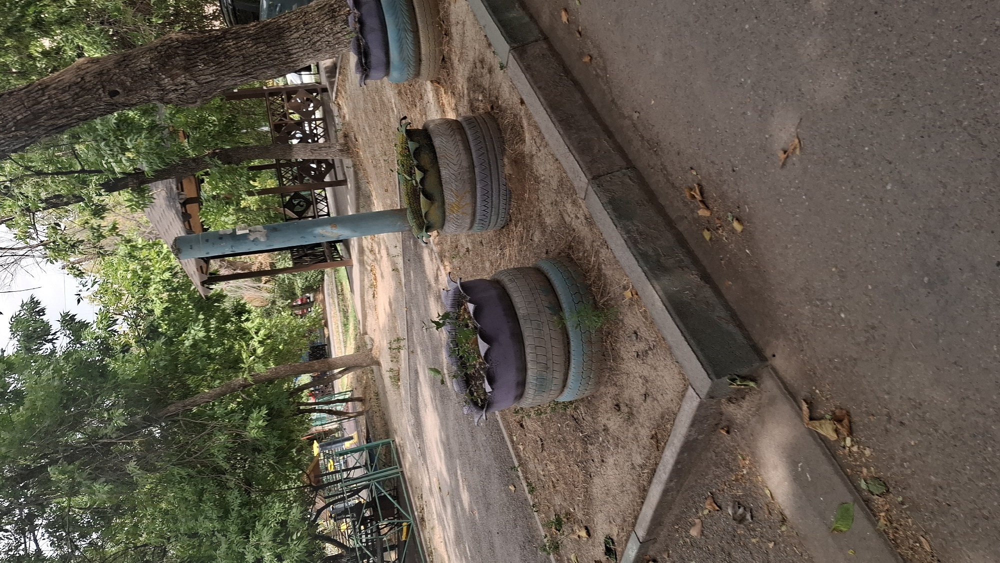 The image depicts an outdoor scene in Shymkent, Kazakhstan, likely a park or recreational area. In the foreground, two planters made from stacked tires are visible. The planter on the left is composed of three tires, painted purple, light blue, and grey, and contains a few small green plants. The planter on the right is slightly taller, with four tires stacked, painted purple, light blue, light grey, and white, and appears to have some greenery or perhaps artificial plants. A light blue cylindrical pole stands between the two planters. The ground around the planters is covered in dry soil and scattered leaves.
In the middle ground, a wooden gazebo with a dark roof and intricate lattice work on its sides is situated amongst trees. The surrounding area shows a paved pathway, more trees with lush green foliage, and what appears to be playground equipment in the background, characterized by green and yellow structures. The overall environment suggests a public space maintained for leisure. The lighting indicates it is daytime, with shadows cast by the trees and structures. The presence of mature trees and the style of the gazebo suggest this is an established area.