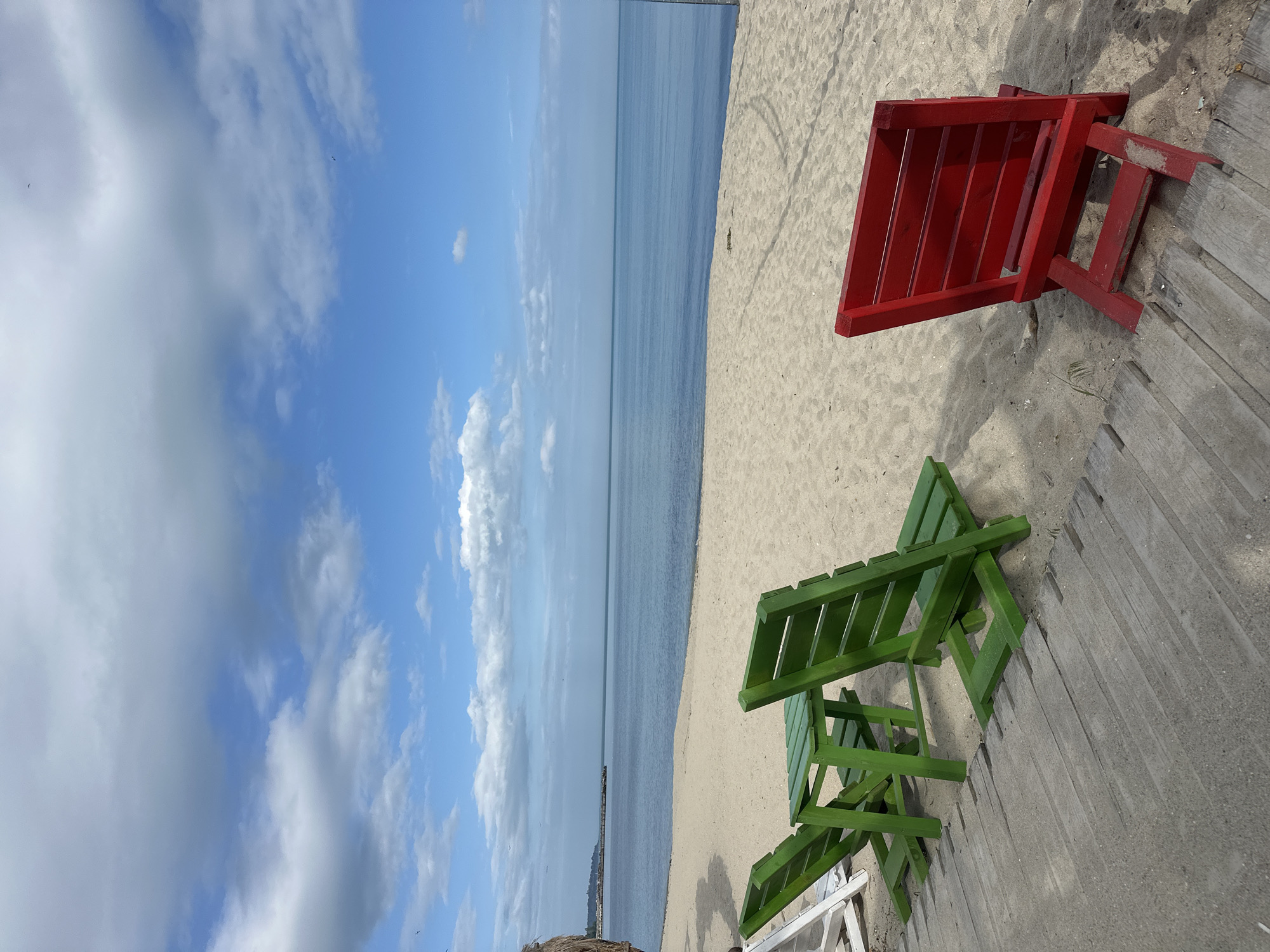 The image is a rotated, high-angle shot of a beach scene with two brightly colored, wooden chairs positioned on the sand next to a wooden walkway. The beach appears to stretch towards a calm body of water that meets a horizon line under a partly cloudy blue sky.
The chairs are the focal point, with one painted red and the other green. They are oriented towards the beach and water. The sand is pale and granular. A wooden walkway borders the sand.
In the distance, the body of water is a dark blue. There is a distant structure or pier visible in the water.
The sky is blue with scattered white clouds.
The presence of the beach, water, and pier, combined with the location context, indicates this is likely near the coast of the city of Varna, Bulgaria.