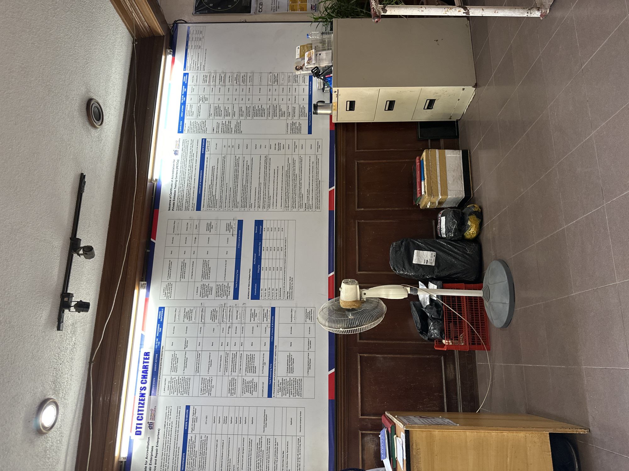 The image depicts an indoor scene within an office space, likely a government service center, possibly located in Davao City, Philippines, as suggested by the "DTI CITIZEN'S CHARTER" prominently displayed on a large informational poster. The poster, which is a central focus, details the procedures and requirements for obtaining an accreditation or certificate, specifically mentioning "Issuance of Certificate of Accreditation for Private Emission Testing Centers (PETCs)" and "Issuance of Certificate of Accreditation for Repair Enterprises."
The room has tiled flooring in a muted brown tone. The walls are paneled with dark wood. In the foreground, on the left, is a wooden counter with a filing cabinet beside it. To the right of the counter, a white oscillating fan stands on a pedestal. Behind the fan, a red plastic crate holds black plastic bags, suggesting discarded or waiting items. Further to the right, stacks of boxed items and rolled-up materials are piled up, and next to them is a tall, beige metal filing cabinet with three drawers.
The lighting appears to be a combination of overhead fluorescent lights and recessed ceiling lights. On the far right edge of the frame, a portion of a white pipe or pole is visible. The overall impression is a functional, somewhat cluttered office environment focused on public service information dissemination. No people are visible in the image.