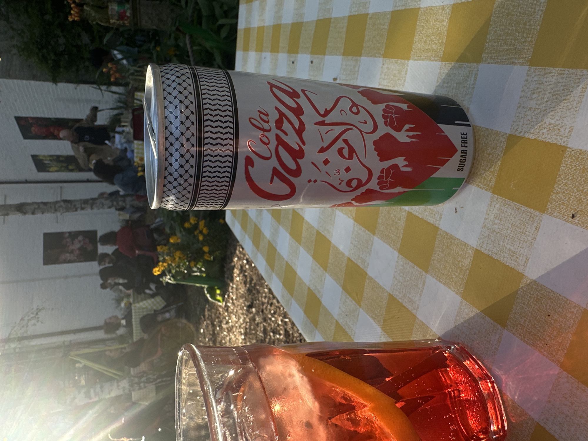 Here's a concise description of the image:
The image shows a can of "Cola Gaza Sugar Free" on a yellow and white checkered tablecloth. The can has Arabic writing and images of raised fists, colored in red, white, and green, likely representing the Palestinian flag. A glass containing a red drink with a slice of citrus is in the foreground. In the background, blurry figures are visible, suggesting an outdoor gathering. The background also includes a building with artwork and foliage. The location is potentially London, United Kingdom.