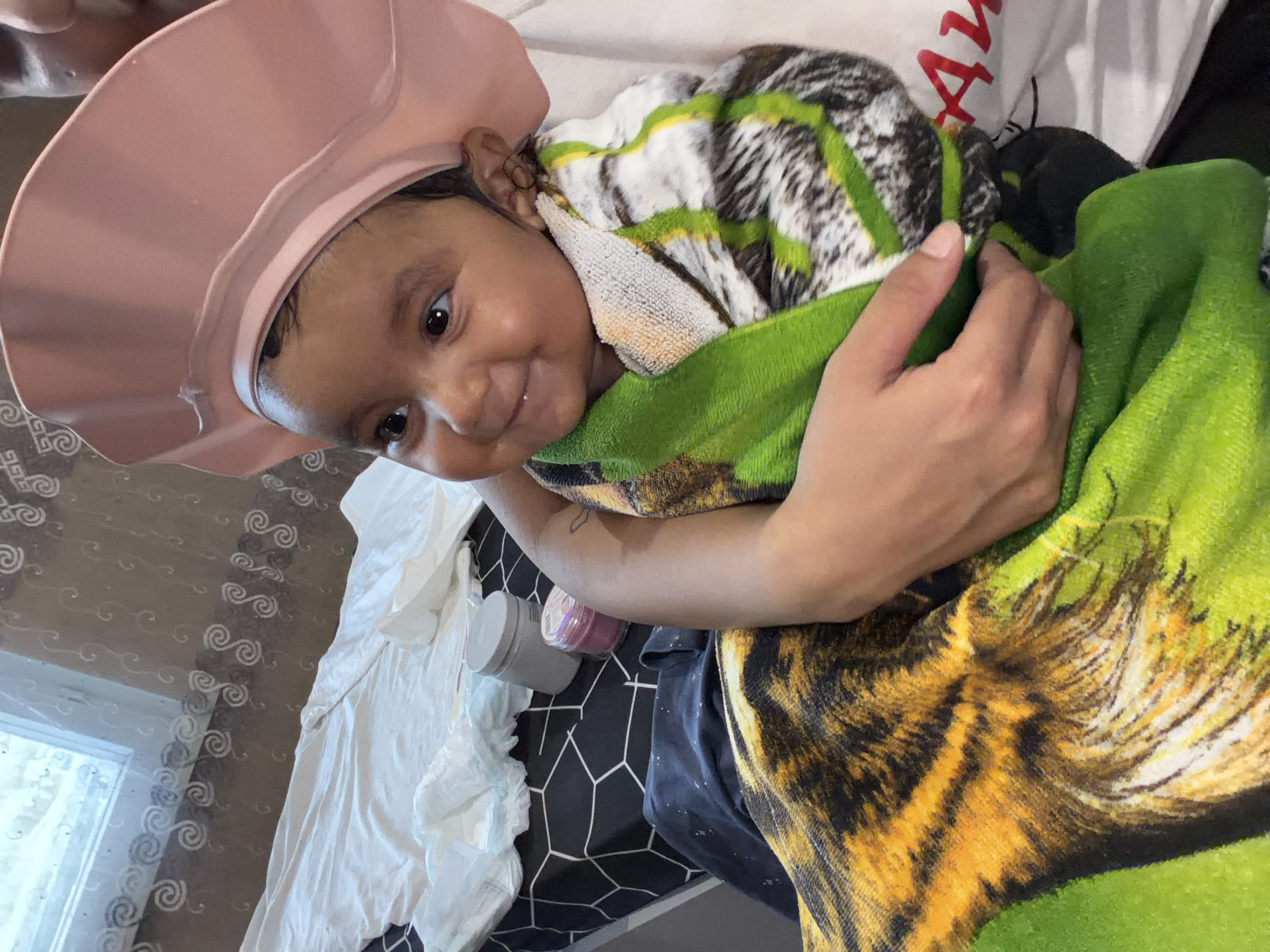 The image shows a baby with dark hair and eyes wearing a pink shower cap and wrapped in towels with a tiger pattern. The baby is being held by an adult whose arm is visible. In the background, there is a bed with white bedding and some containers. A window with patterned curtains is also visible. The scene appears to be indoors. The text "AWA" is partially visible on the adult's shirt. The specific location of Razhevo Konare, Bulgaria, is not discernible from the visual information in the image.