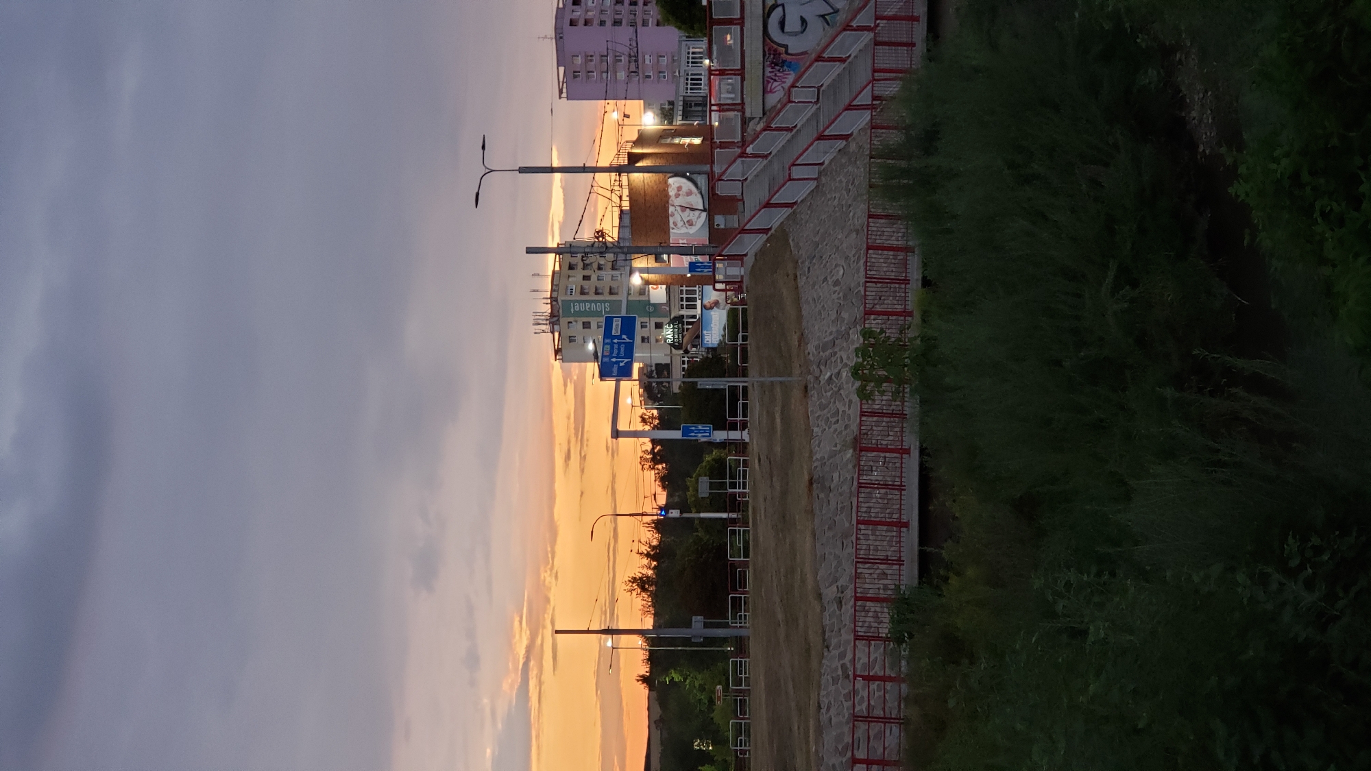 This image captures a sunset scene in Prešov, Slovakia. In the foreground, dense green vegetation obscures a body of water. A red-railed bridge spans across the middle ground, with a stone embankment supporting it. Beyond the bridge, an elevated walkway with red railings leads upwards towards buildings. In the background, several multi-story buildings are visible, including a light-colored building with the Slovak flag and text "Slovakiell" on it, and a pink apartment building. Streetlights and traffic signs are present along the road. The sky is a gradient of dark clouds and the warm orange and yellow hues of a sunset.