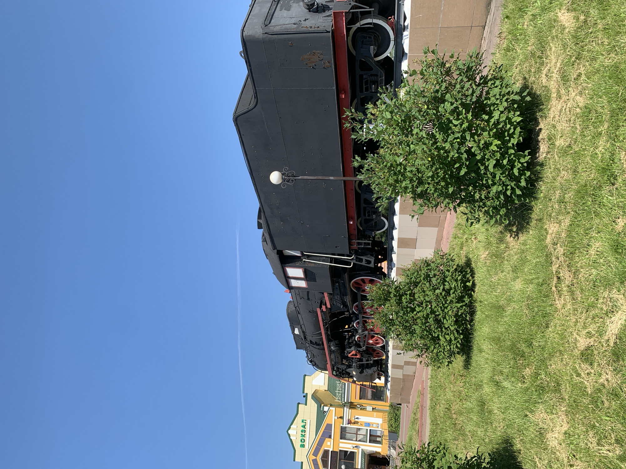 An outdoor, eye-level, wide shot captures a restored black steam locomotive on display in front of a yellow and green building labeled "ВОКЗАЛ" (VOKZAL, meaning "station" in Russian) under a clear blue sky. The locomotive, with its large red-rimmed wheels, is positioned on a raised platform resembling train tracks, with several well-maintained green bushes and mowed grass in the foreground. The building, likely a train station, exhibits a classic architectural style with arched windows and a prominent green roof. The overall scene suggests a sunny day in Prokopyevsk, Russia, where the locomotive serves as a historical monument, likely at the city's railway station. There are no people or animals visible in the frame.