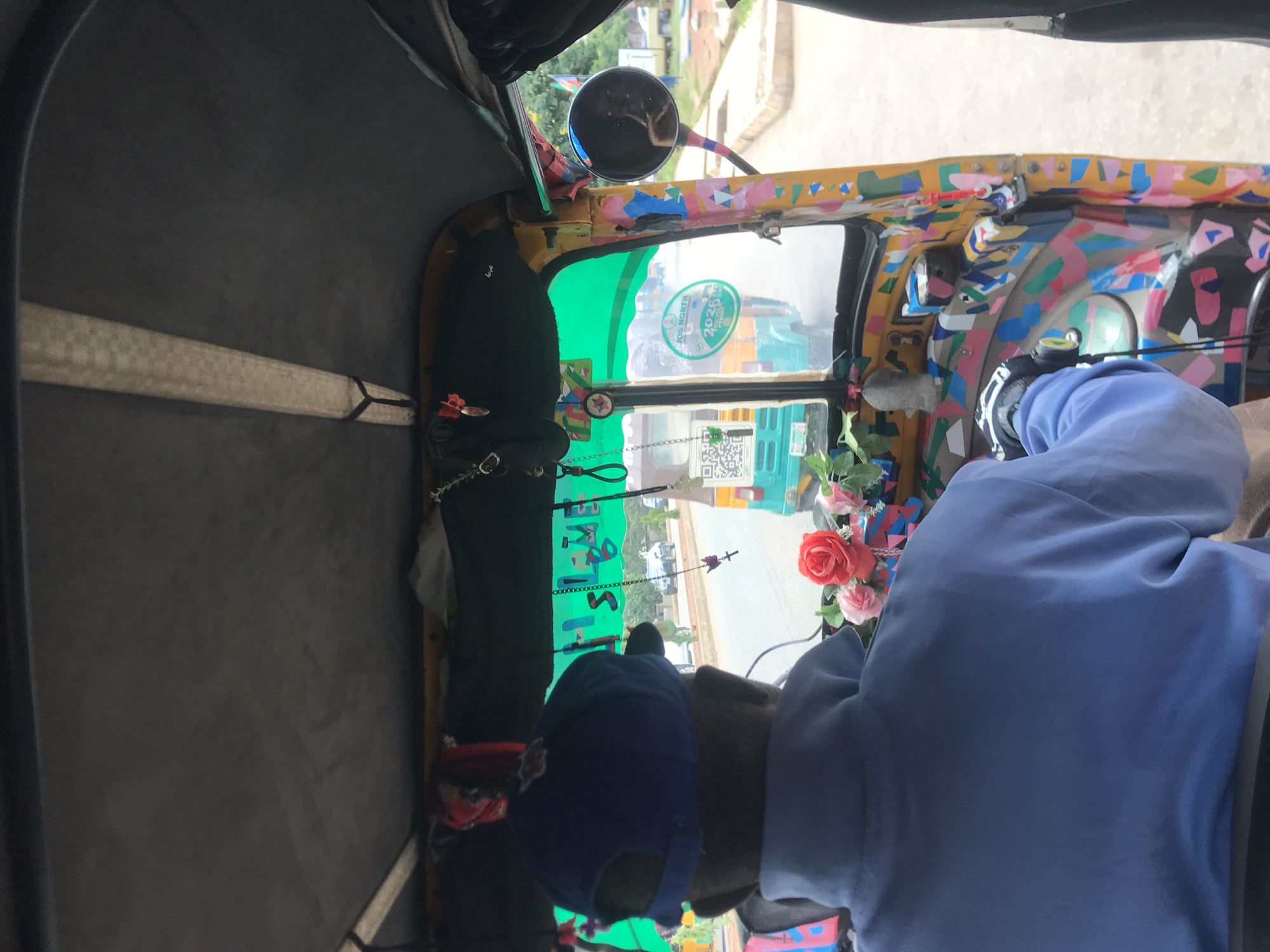 The image shows the interior of a decorated auto-rickshaw, commonly known as an "abajaj" in Nigeria, in Jos, Nigeria. The driver, wearing a blue baseball cap and a light blue shirt, is visible from the back as they operate the vehicle. The auto-rickshaw is brightly painted with a variety of colorful geometric shapes in pink, blue, yellow, and black. There are decorative items hanging from the interior, including a bunch of artificial flowers with red and pink roses, a small black cross, and a beaded chain. A side mirror on the right reflects a distorted image of the interior. The exterior of other auto-rickshaws and some buildings are visible through the windshield. A sticker on the windshield reads "JOS NORTH 2026 ELECTION". The overall impression is one of vibrant, personalized transportation within the city.