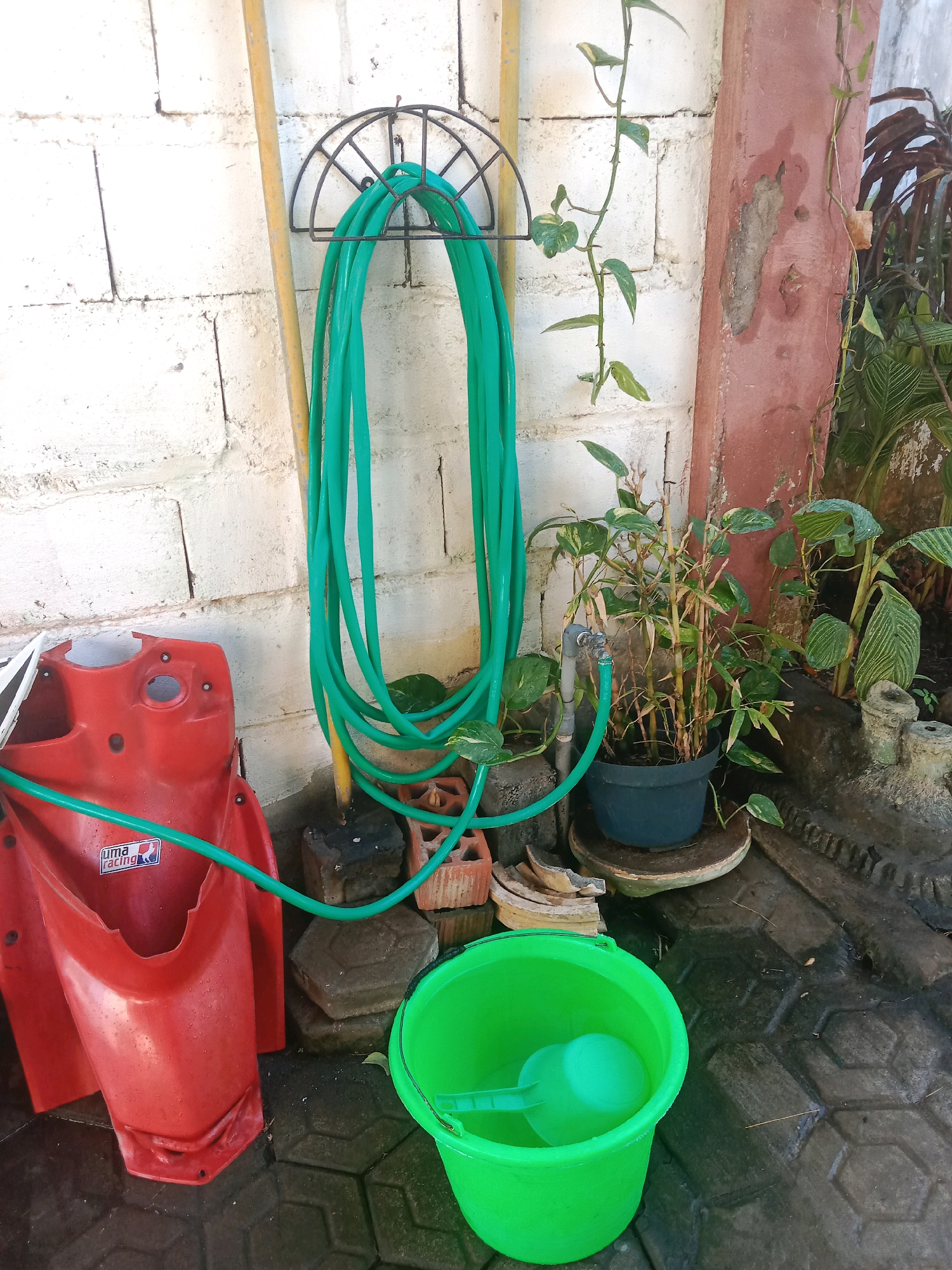 This image depicts an outdoor scene in the city of Lemahkembar, Indonesia, likely a backyard or a utility area. A bright green garden hose is coiled on a black metal holder attached to a white brick wall. To the left of the hose, a yellow pole stands vertically. In front of the wall, a red plastic fender-like object, possibly from a motorcycle or scooter, is placed on the ground. A green hose snakes across the ground, connected to a spigot on a potted bamboo plant. The plant is in a dark blue pot, which sits on a circular weathered concrete base. To the right of the bamboo plant, other greenery is visible. In the foreground, a vibrant green plastic bucket with a matching scoop sits on a patterned grey paved surface. The ground appears wet, suggesting recent use of water or rain. The overall impression is one of a functional outdoor space, perhaps used for gardening or cleaning.