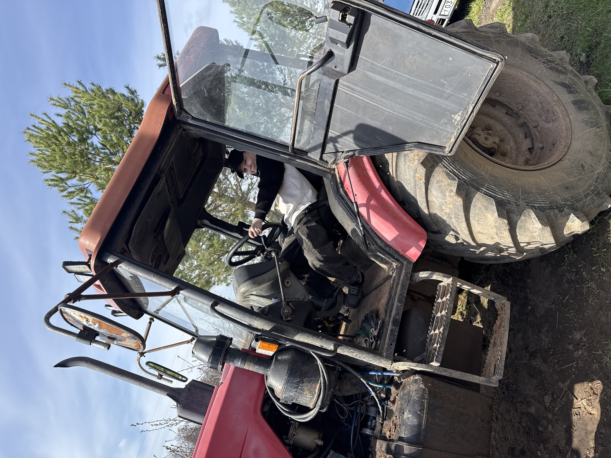 A young person is sitting in the driver's seat of a large red and black tractor. The tractor is parked outdoors on what appears to be a grassy, possibly muddy, surface. The tractor's door is open, revealing part of the interior. The sky is visible, suggesting it is daytime. The photo was taken in Уральское, Russia.
