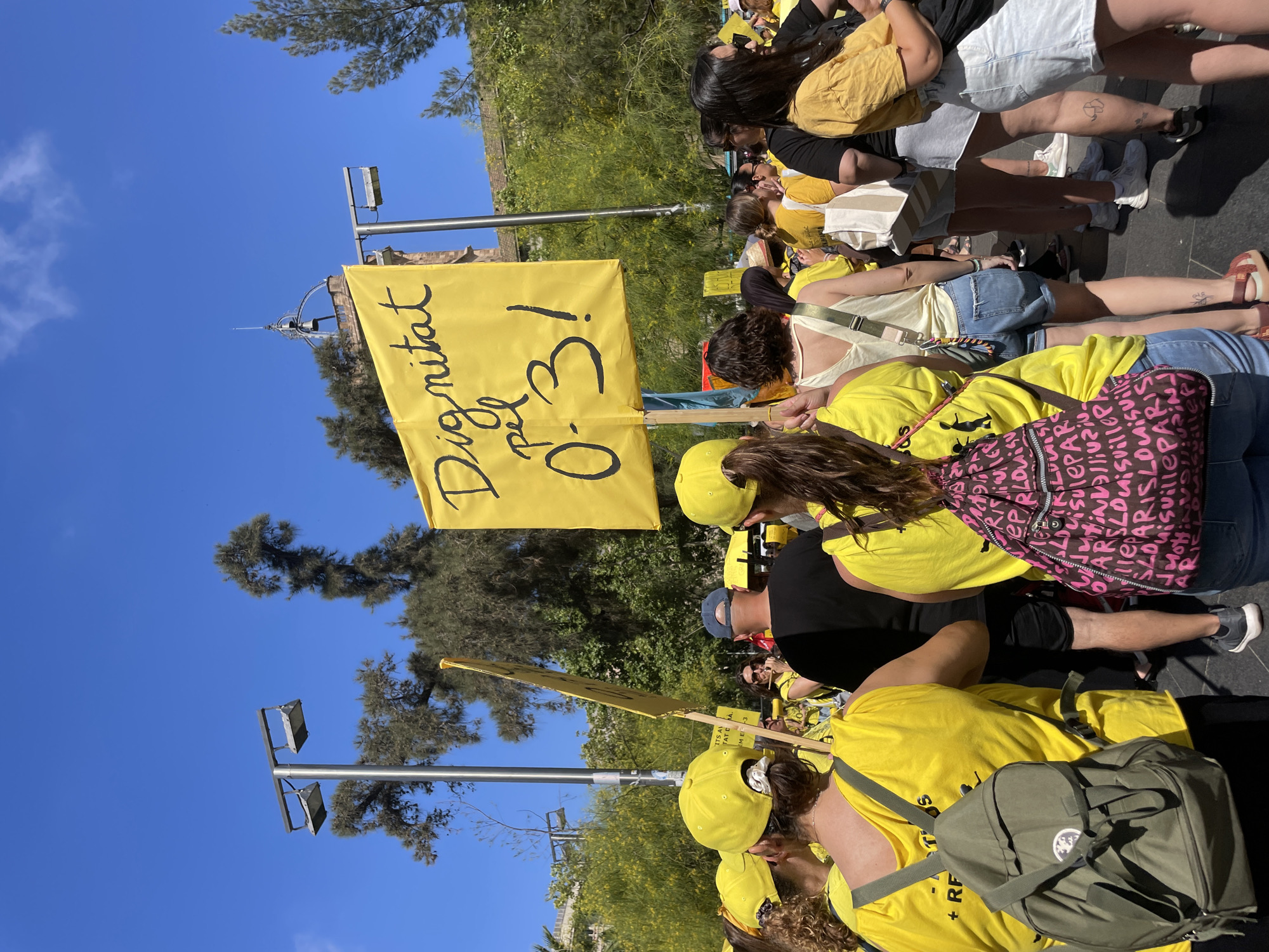 A group of people are gathered outdoors on a sunny day. They are wearing yellow shirts and caps, and many have yellow banners. One banner in the foreground has black handwritten text that reads "Dignitat pel 0-3!". The crowd appears to be participating in a protest or demonstration. In the background, trees and a blue sky are visible. The environment suggests an urban setting, likely a public square or street in Barcelona, Spain, given the Catalan text on the banner. The people are diverse in age and gender, and their focus is directed forward, towards the front of the gathering.