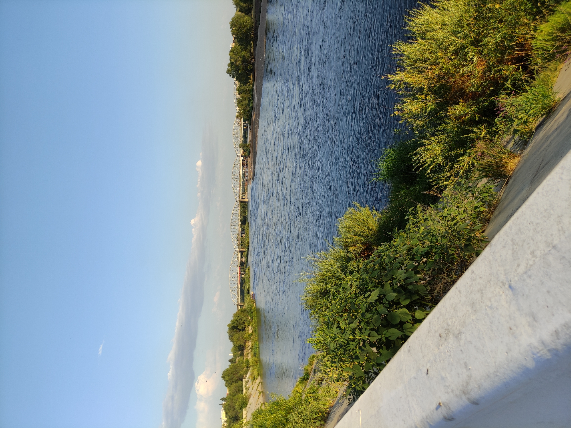 The image depicts a wide, calm river flowing under a clear blue sky with scattered white clouds. A long, multi-span bridge crosses the river in the distance, with a small boat visible on the water in front of it. The riverbanks are lined with lush green vegetation, including trees and bushes. In the foreground, a concrete barrier runs along the edge of what appears to be a promenade or embankment, providing a vantage point over the river. The scene suggests a peaceful, natural setting within an urban environment. This is likely a view of the Irtysh River in the city of Semey, Kazakhstan, given the presence of a prominent bridge and the general landscape.