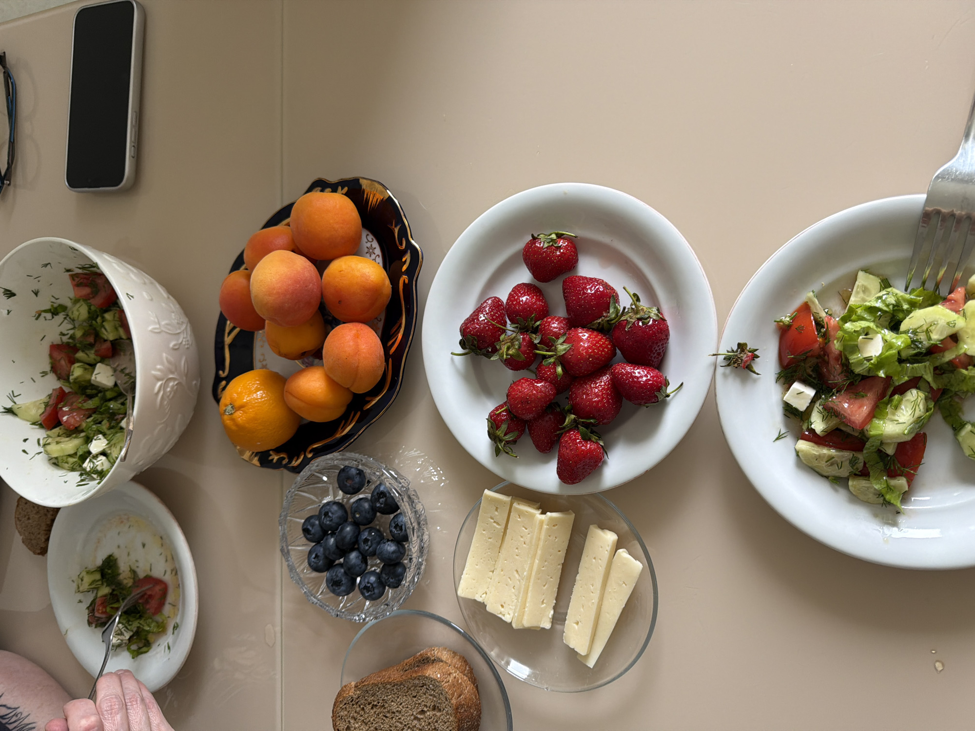 The image shows a beige tabletop with various food items arranged on it. There are two bowls of salad, one large and one small, containing tomatoes, cucumbers, feta cheese, and dill. A decorative plate holds apricots and one orange. A white plate is filled with strawberries. A crystal bowl contains blueberries. Another glass bowl holds slices of bread. A plate displays slices of cheese. A fork is visible in the salad on one of the plates. A mobile phone and glasses are on the top left side of the scene. There are human hands holding the fork. The location, Izhevsk, Russia, cannot be confirmed from the visual information.