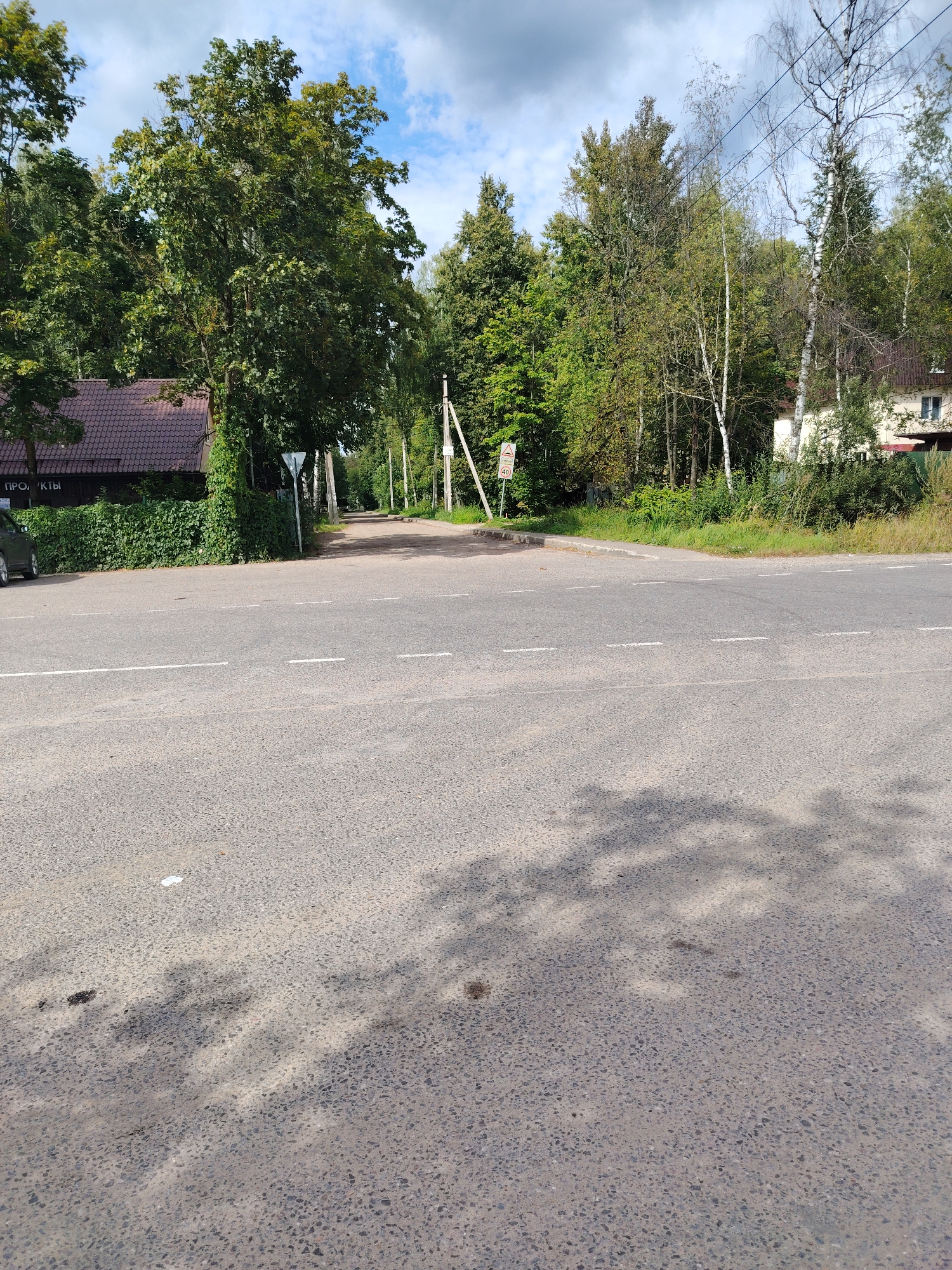 The image shows a wide, asphalt road intersection in a rural or suburban area. In the foreground, the road is marked with white dashed lines. The background features a dense green treeline with a mixture of deciduous and coniferous trees. On the left side of the intersection, there is a building with a dark, patterned roof, partially obscured by dense green bushes. A dark-colored car is parked to the left of this building. Further down the road, several utility poles are visible, and in the distance, a building with a light-colored facade and red roof can be seen. A triangular road sign with a speed limit of 40 is also visible on the left. The sky is partly cloudy with patches of blue. The overall impression is a quiet, somewhat undeveloped intersection on a sunny day.