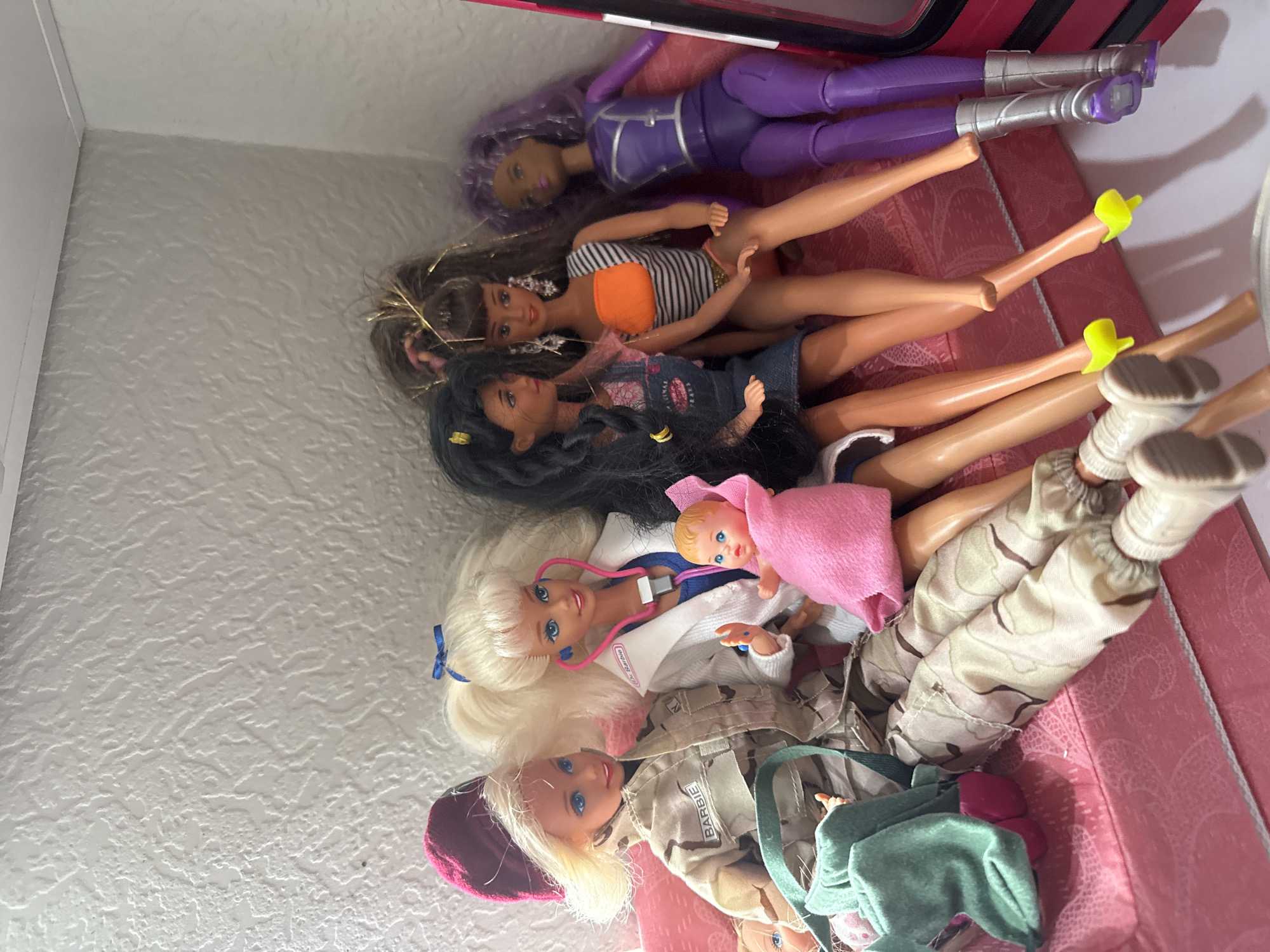 A group of five Barbie dolls and one baby doll are posed on a pink sofa against a textured white wall. From left to right, the first Barbie doll wears a camouflage uniform and a maroon beret, with a green bag on her lap. The second Barbie doll is dressed as a doctor with a white coat and a pink stethoscope. Next to her is a baby doll wrapped in a pink blanket. The third Barbie doll has dark hair and wears a denim overalls dress. The fourth Barbie doll sports teased brown hair and a striped orange and white top with a pink skirt. The last Barbie doll has purple hair and wears a purple jumpsuit with silver boots. The lighting is bright, casting shadows on the dolls and the wall. The scene appears to be indoors, in a domestic setting, without any specific urban indicators to suggest a city or location.