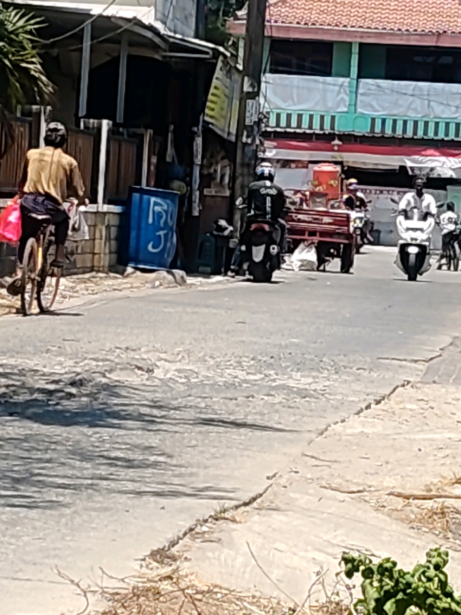 In Mekarmukti, Indonesia, a street scene unfolds under a bright sky, capturing a moment in the day. A person on a bicycle, dressed in a yellow shirt and dark pants, pedals down a paved road. A red bag hangs from the bicycle. To the left of the cyclist, a blue drum with white lettering is visible. Further down the road, several motorcycles are present. One motorcycle, ridden by a person wearing a black jacket and white helmet, is in the foreground. Another motorcycle, ridden by a person in a white shirt, is further to the right. Behind them, a red cart is being pulled by a motorcycle. Buildings line the street, some with tiled roofs and patterned awnings. The road surface appears worn and cracked in places, with a lighter colored, rougher surface visible on the right side of the frame. Green foliage is present in the lower right corner of the image.