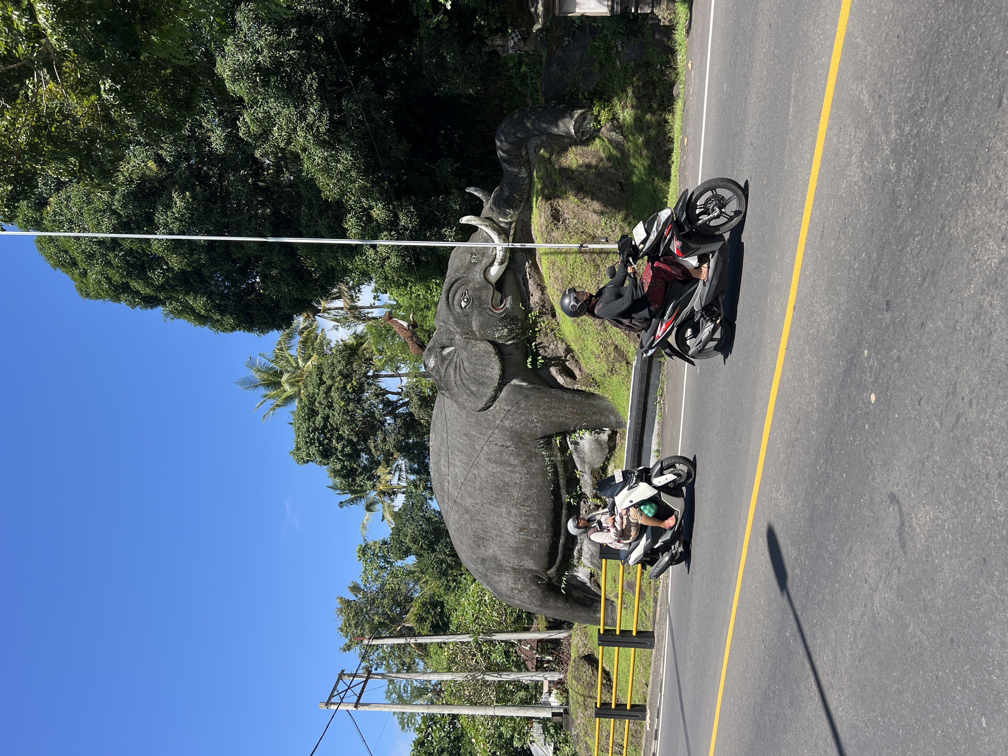 The image shows a large elephant statue on the side of a road in Banjarangkan, Indonesia. Two people are on motorcycles parked near the statue. The road has a yellow center line. The sky is blue, and there are trees and power lines visible. The statue is grey and appears to be made of stone or concrete.