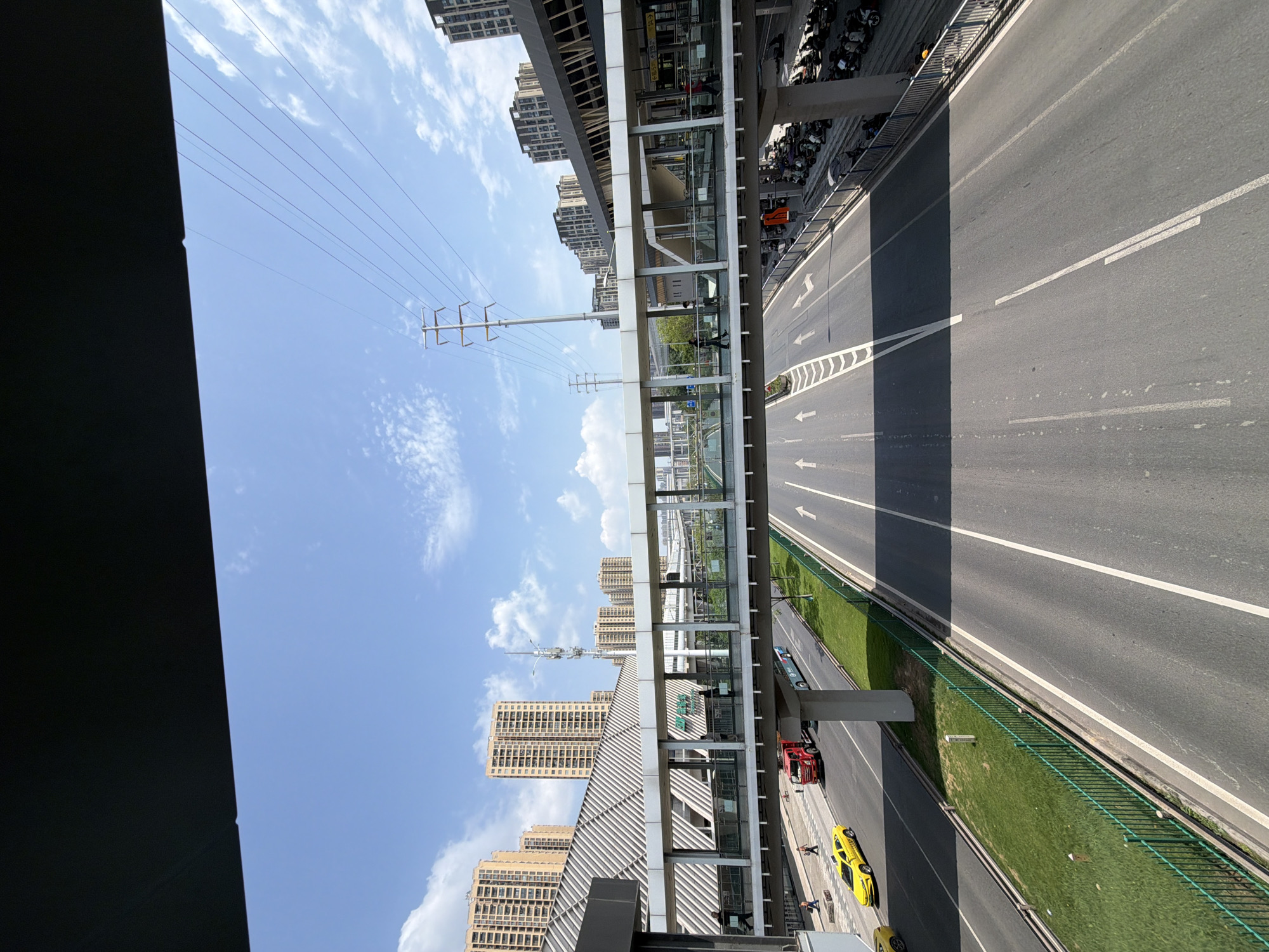 The image depicts a view from a pedestrian overpass in Tiaodeng, China, looking down onto a multi-lane road with traffic. The overpass itself is a modern structure with glass walls, and several pedestrians are visible walking across it. Below, on the road, a yellow taxi, a red truck, and a bus are traveling. The road is well-maintained with clear lane markings and arrows indicating direction. On the sides of the road, there are grassy verges and a green fence. In the background, tall residential buildings and modern urban architecture dominate the skyline under a bright blue sky with scattered clouds. Power lines and utility poles are also visible, along with parked motorcycles on the right side of the image.