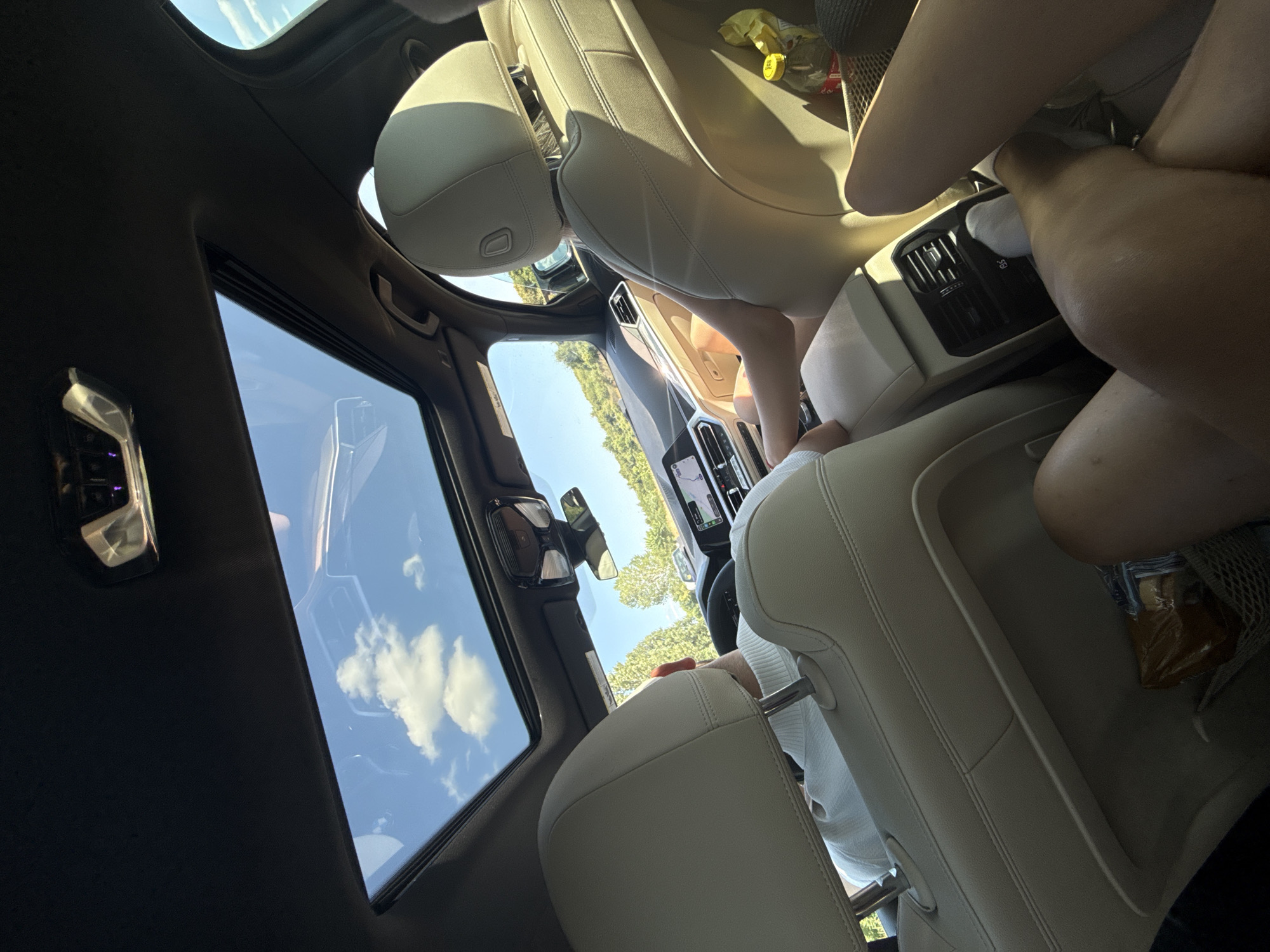 The image, taken from inside a car with a panoramic sunroof, shows the interior of the vehicle from the perspective of the rear passenger seats. The sunroof reveals a bright blue sky with scattered white clouds. The seats are upholstered in a light-colored leather. In the front, a person wearing a white sweater is visible from the back, seated in the driver's seat and looking forward. The car's dashboard and navigation screen are also visible. In the foreground, the legs of at least two individuals are visible in the rear seats, suggesting multiple occupants. A mesh storage bag is attached to the back of one of the front seats, containing a packaged item. Another passenger's legs are visible in the right rear seat, next to a yellow bag and a bottle with a red cap. The ambient light suggests it is daytime. The landscape visible through the sunroof and side windows appears to be rural, with trees and greenery. This scene is not set in the city of Boiștea de Jos, Romania, as there are no identifying landmarks or contextual clues to support this location.
