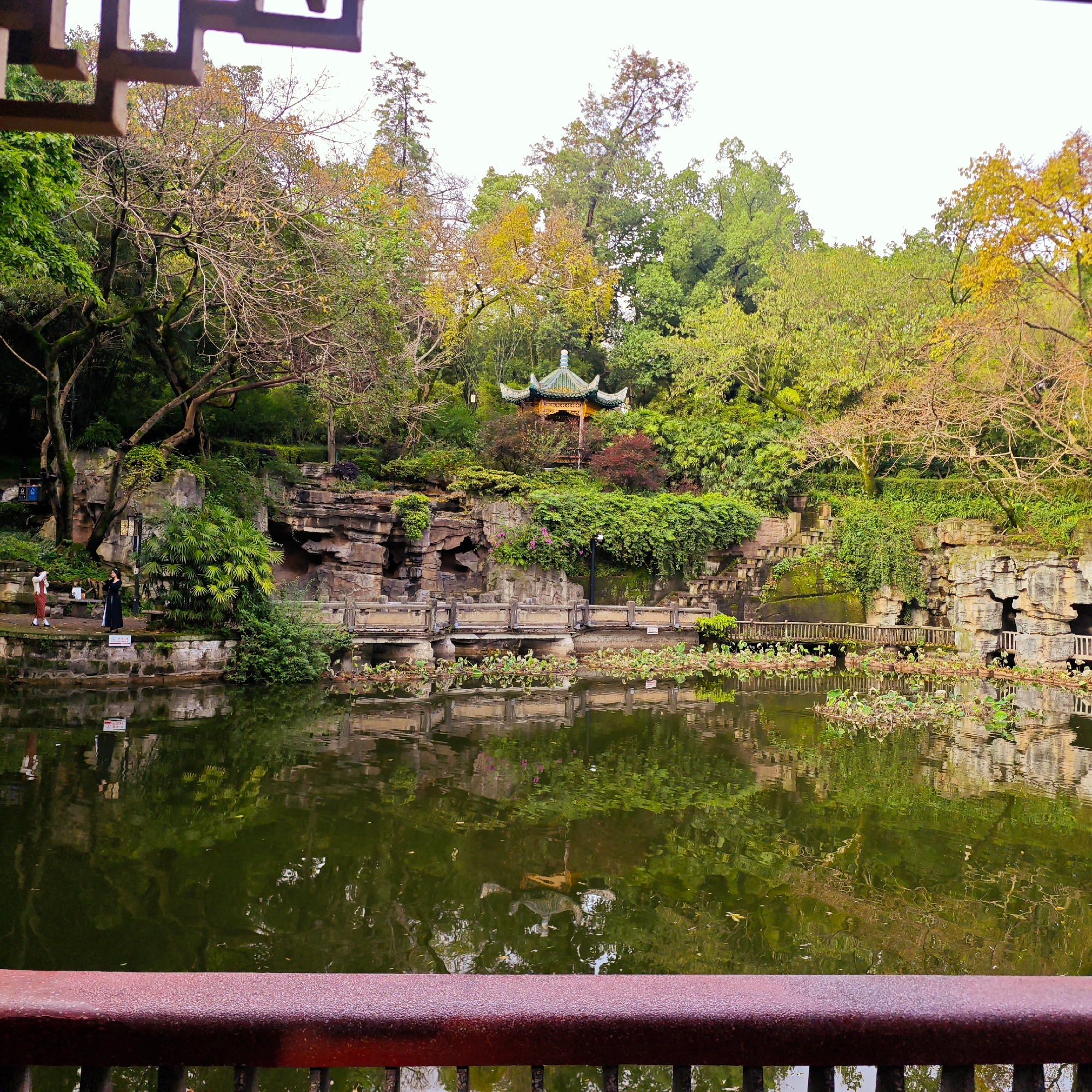This media file presents a serene landscape within a traditional Chinese garden, likely situated in Yuzhong District, China. The scene is dominated by a calm, dark green pond or lake in the foreground and midground, its surface reflecting the overcast sky and lush vegetation. Patches of water lilies or similar aquatic plants are visible on the water's surface. A rusty red metal railing, part of a bridge or viewing platform, frames the bottom of the image.
The far bank features layered stone retaining walls and rock formations, heavily adorned with dense green foliage, moss, and bushes. Several rustic wooden or stone bridges extend over sections of the water along the bank, connecting pathways. A distinctive traditional Chinese pavilion, with a tiered green roof and ornate detailing, is nestled among the trees on a verdant hillside in the background. The trees exhibit a mix of vibrant green, autumnal yellow, and bare branches, suggesting a season like late autumn.
A few people are visible on the left side of the far bank. One individual is dressed in a long, dark traditional Chinese garment, possibly a Hanfu, standing beside another person in lighter attire. They appear to be enjoying the park, perhaps sightseeing or taking photographs. The sky is bright but overcast, indicating daytime with soft, diffused lighting. Although reflected signs in the water are unreadable, the overall atmosphere conveys tranquility and natural beauty, characteristic of a well-maintained Chinese garden.