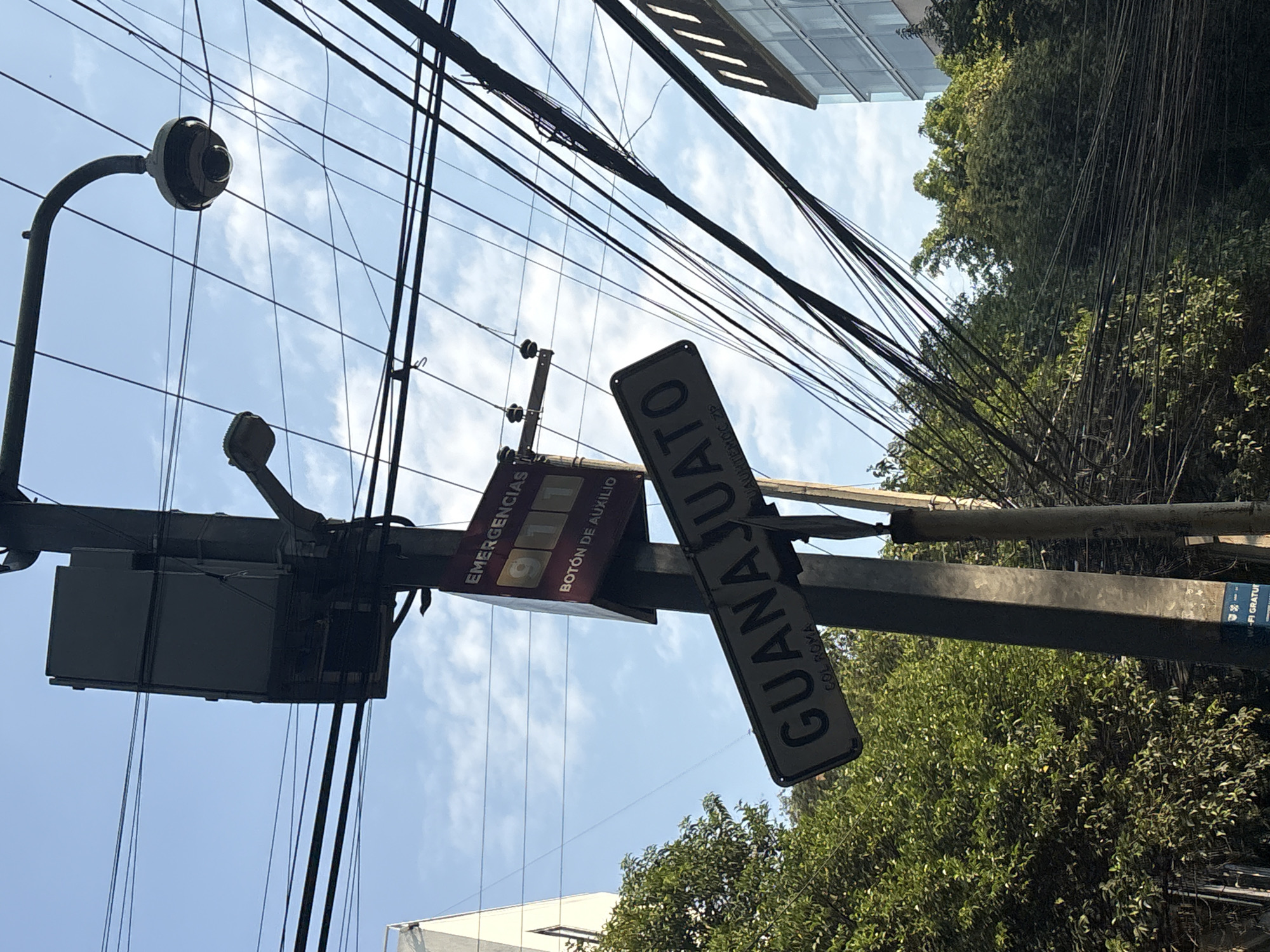 The image shows a street scene in Mexico City, Mexico. The view is looking up at a street sign that reads "GUANAJUATO COL. ROMA" attached to a utility pole. Next to the street sign is a sign for emergencies, showing "911" and "BOTON DE AUXILIO". There are numerous electrical wires overhead and a security camera is mounted on the utility pole. Trees and a building are visible in the background.
