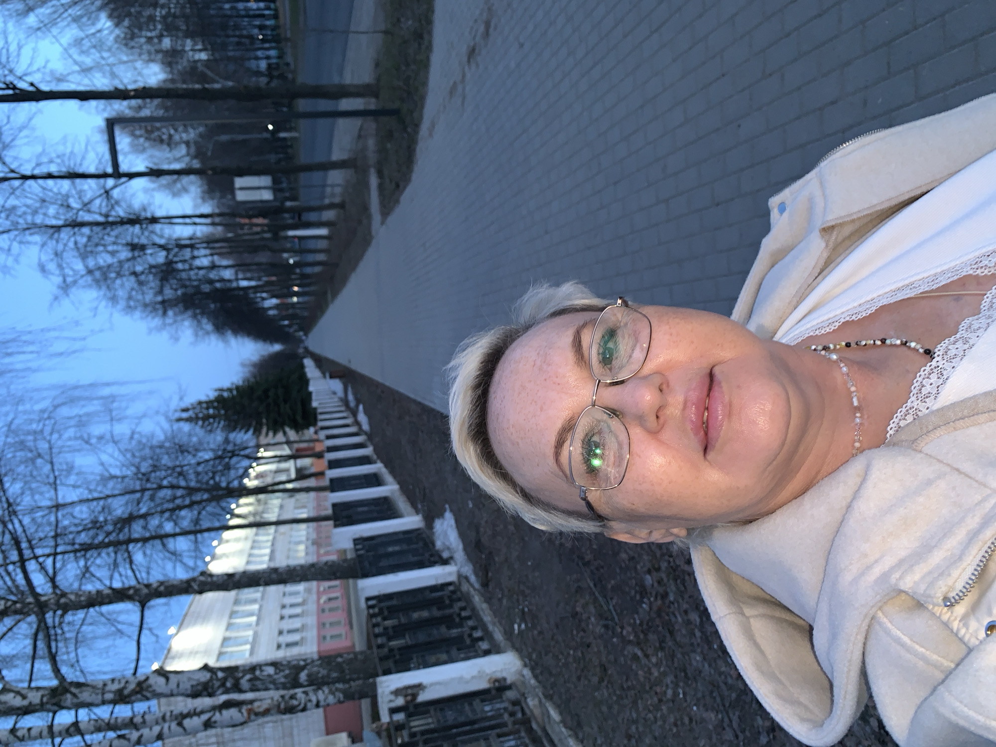 The image is an outdoor selfie featuring a woman in the foreground, with a long path and buildings in the background under a twilight sky.
The main subject is a woman with short, light blonde hair and visible freckles on her face. She is wearing gold-rimmed glasses that reflect green light, and she has a gentle smile with slightly parted lips. Her attire includes a creamy-white, possibly fleece or sherpa, zip-up jacket worn over a white top with a delicate lace trim around the neckline. A multi-colored beaded necklace with natural, white, and black tones adorns her neck.
Behind the woman, a long, straight paved path or sidewalk stretches into the distance. It is bordered on both sides by tall, slender deciduous trees with bare branches, suggesting a colder season. On the left side of the path, several multi-story buildings are visible, including a tall apartment-style structure with numerous windows, some of which are illuminated, and sections with pink and grey/white facades. An evergreen tree stands among the bare trees further down the path. The ground surface appears to be asphalt or concrete.
The scene is set during dusk or early evening, characterized by the deep blue sky and the activation of artificial lights on the buildings. The overall ambiance suggests a cool temperature. The woman's expression is calm and pleasant. There is no visible text in the image. The setting is an urban or suburban environment, consistent with the provided location context of Chaikovsky, Russia.
