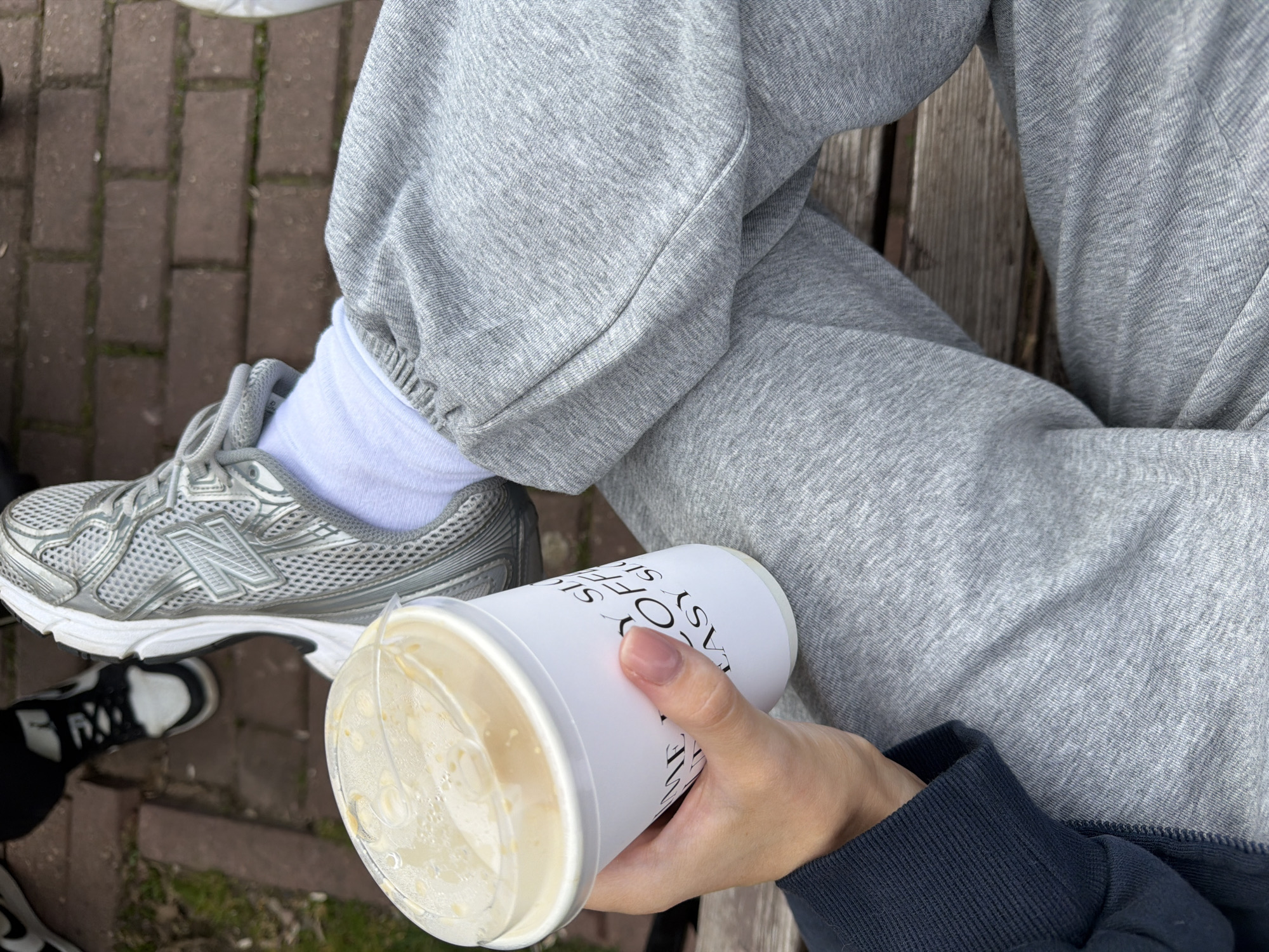The image shows a person sitting outdoors, likely on a bench, holding a disposable cup with a clear lid containing a liquid, possibly a coffee drink. The person is wearing grey sweatpants and white socks. On their feet, they have a silver New Balance sneaker and a partial view of another black and white shoe is visible. The background consists of brick paving and some green vegetation. The location information is not discernible from the image.